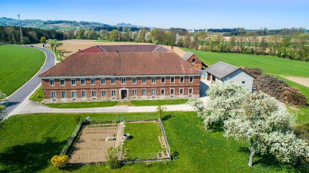 Luftaufnahme eines Bauernhofs mit roten Ziegeldächern, umgeben von  Feldern und blühenden Bäumen.