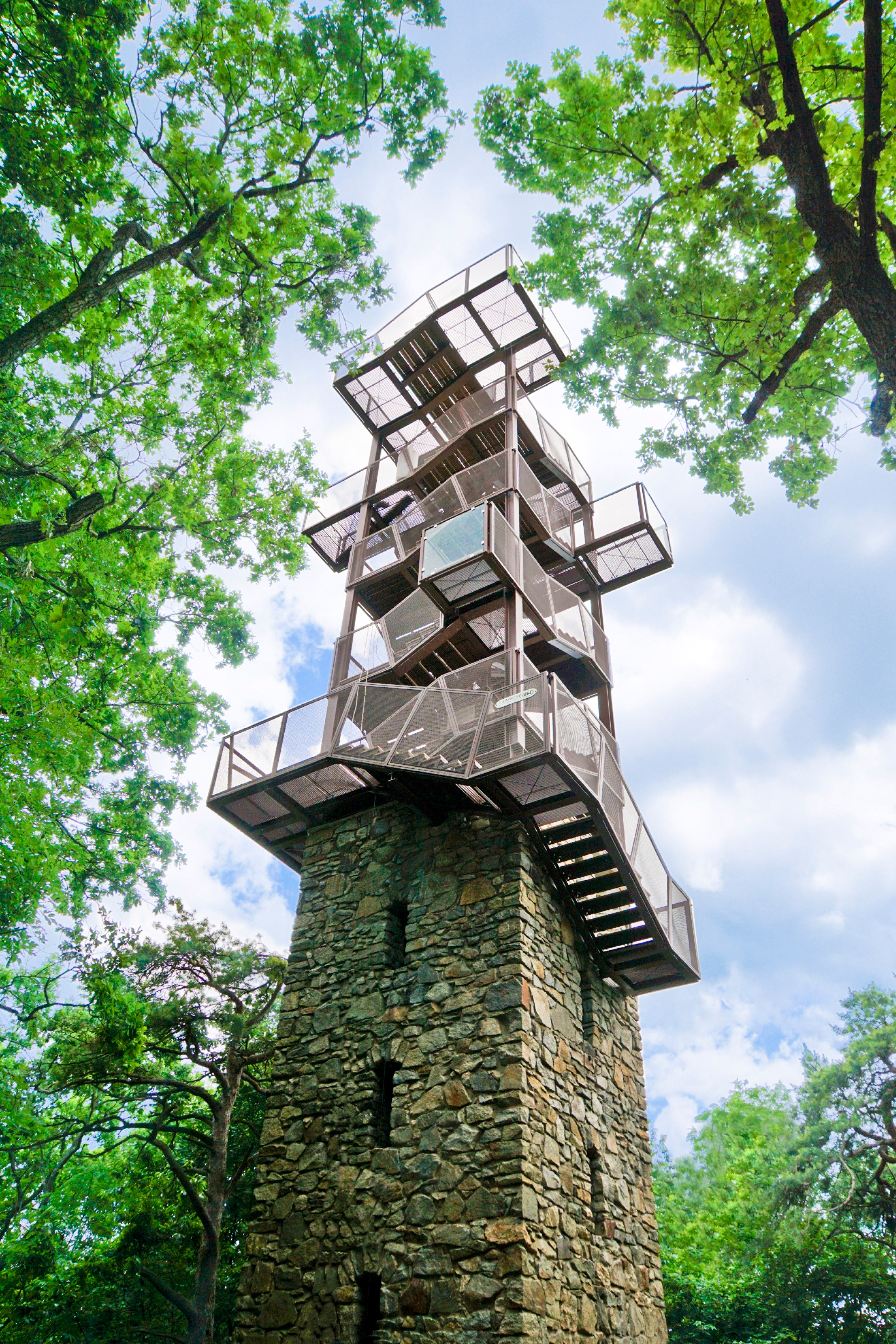 Ein moderner Aussichtsturm aus Metall und Stein, umgeben von grünen Bäumen, ragt in den blauen Himmel.