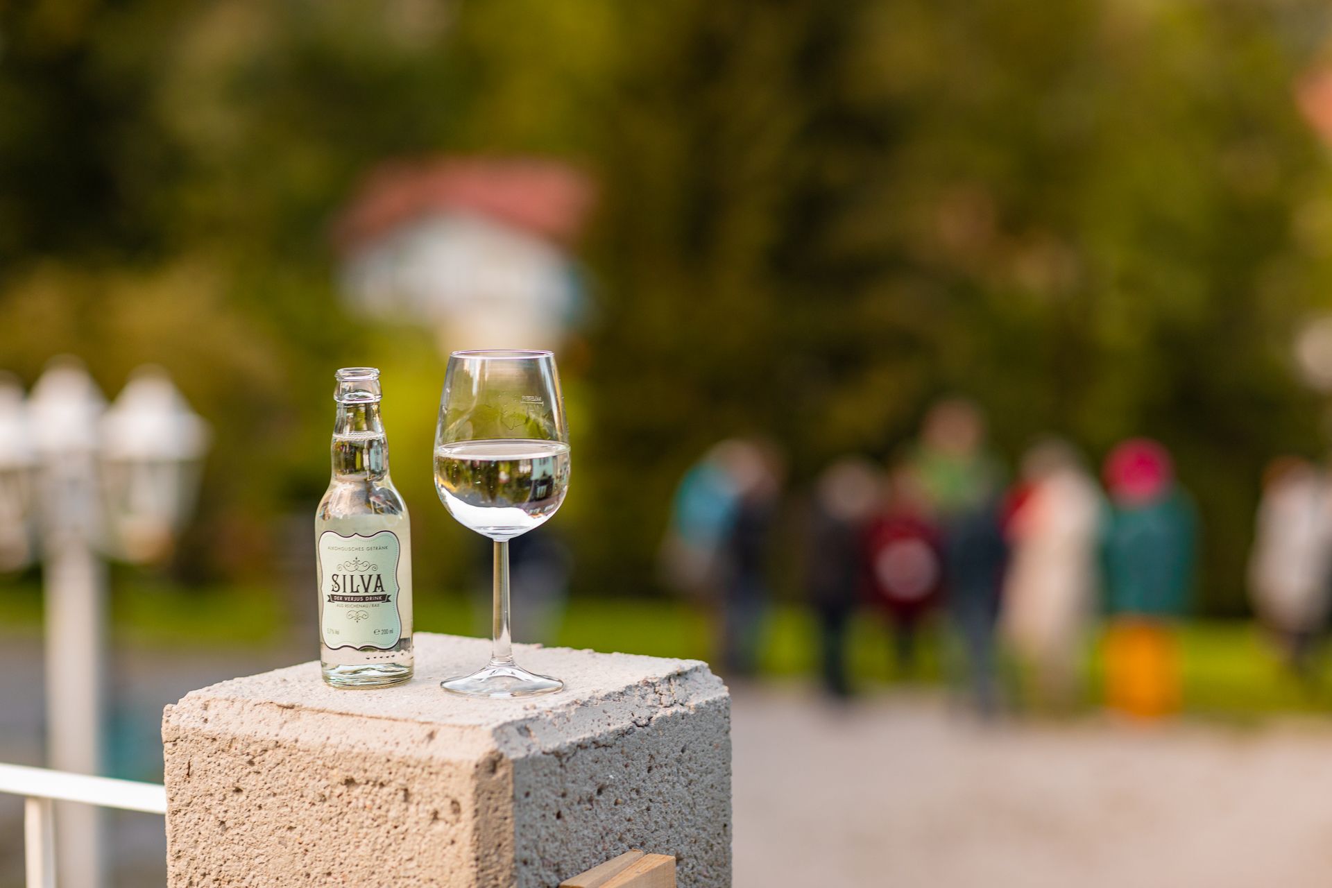 Flasche und Glas mit Silva-Verjus-Drink auf einem Betonpfeiler, unscharfe Personen im Hintergrund.