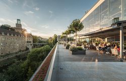 Panorama einer Flusslandschaft mit Schloss und moderner Architektur, umgeben von grünen Hügeln.