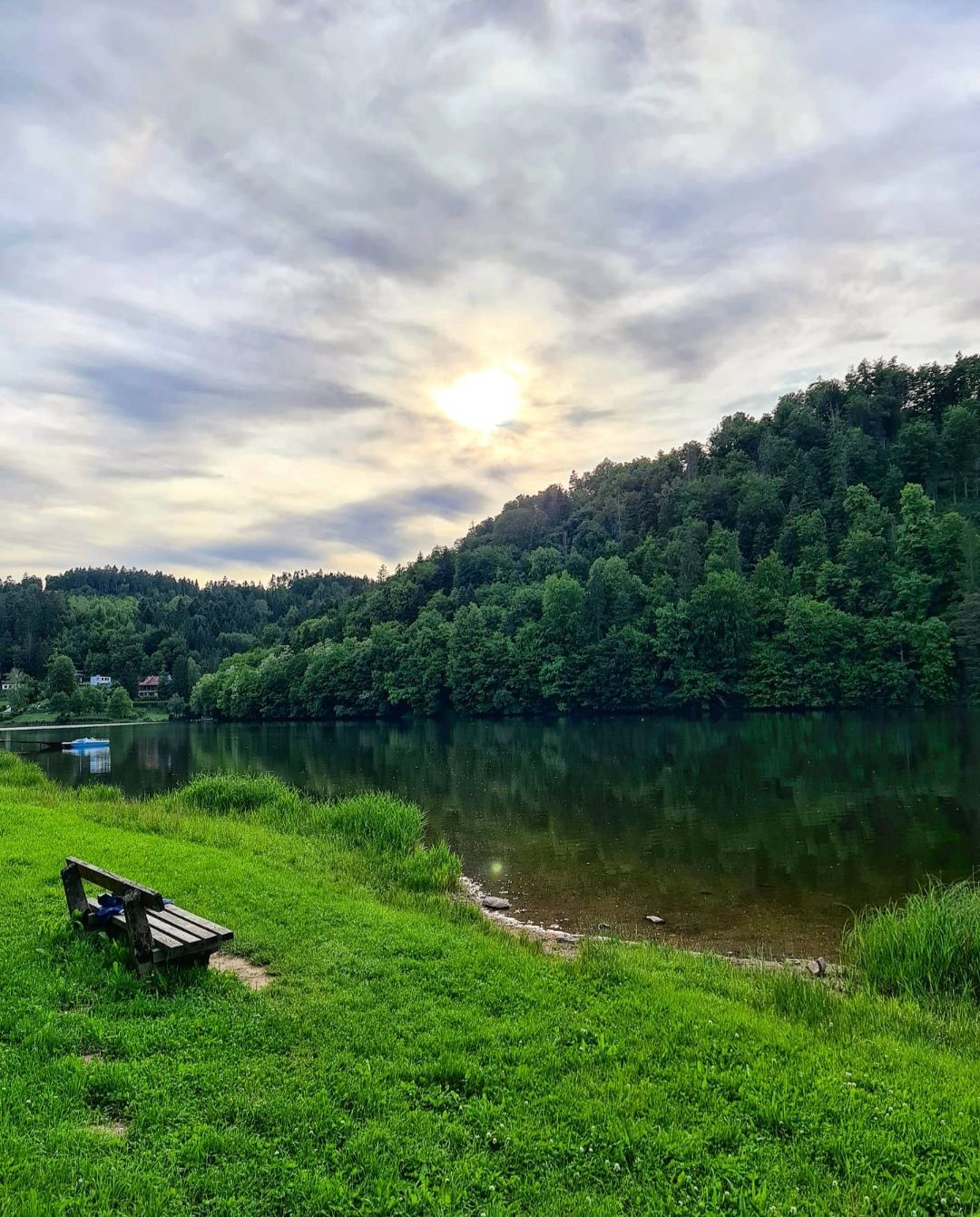 Eine Bank steht auf einer grünen Wiese am Ufer des Stausees Thurnberg, umgeben von bewaldeten Hügeln und unter einem bewölkten Himmel mit sichtbarer Sonne.