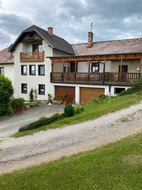 Ein zweistöckiges Haus mit Balkon und Garage, umgeben von grüner Landschaft und einem bewölkten Himmel.