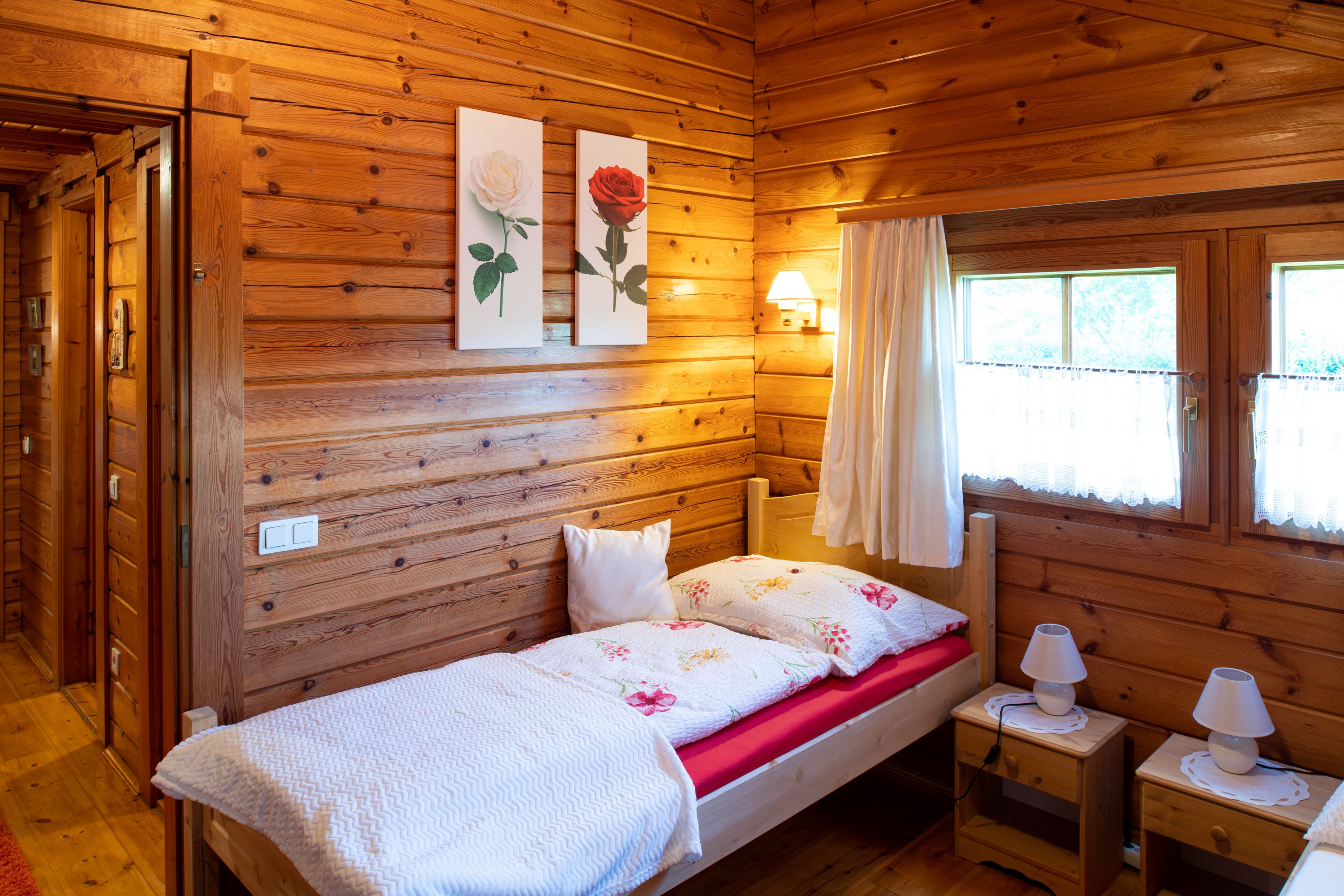 Gemütliches Schlafzimmer mit Holzwänden, Einzelbett, zwei Nachttischen und Fenstern mit Vorhängen.
