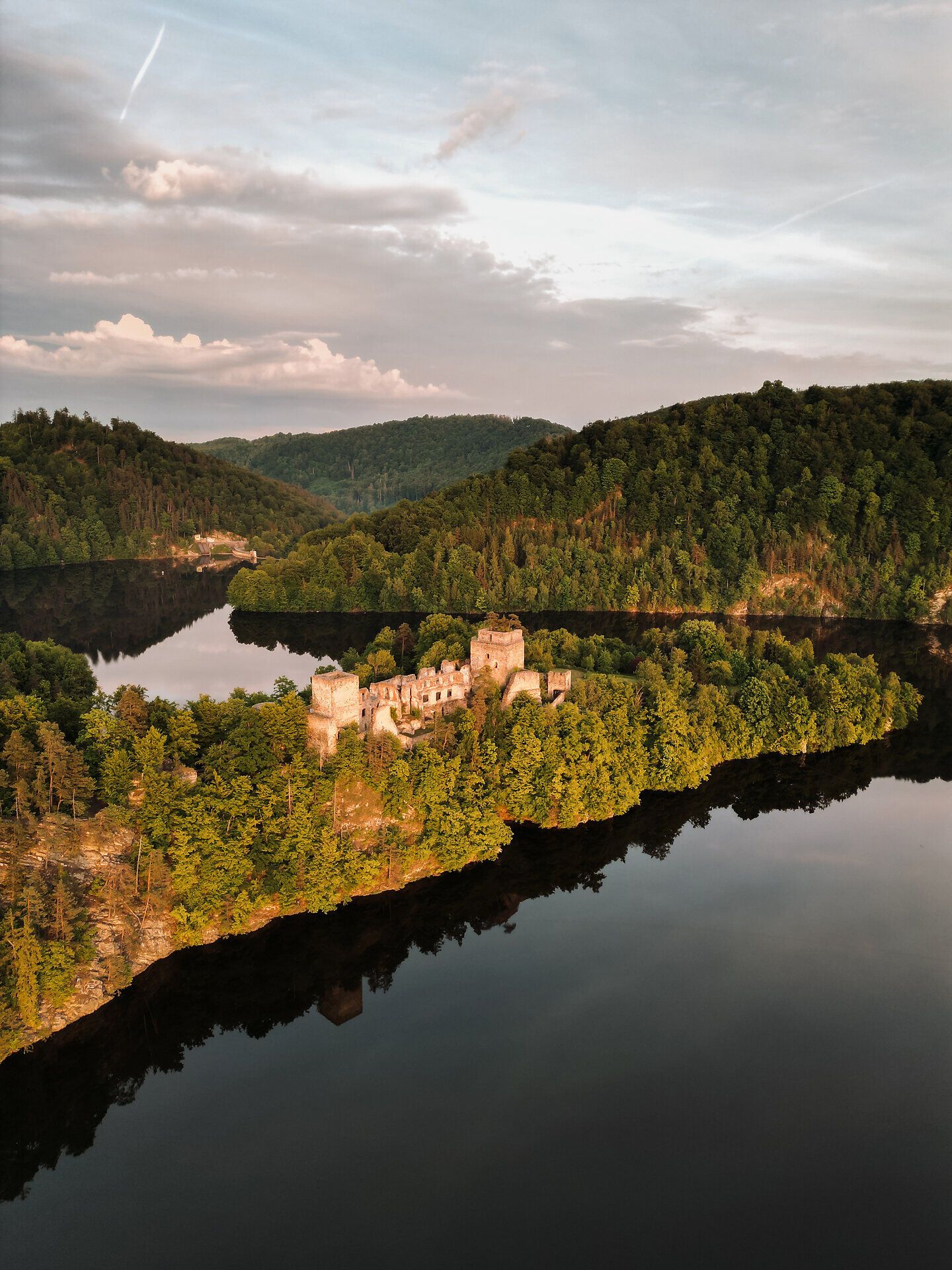Eine Luftaufnahme der Burgruine Dobra im Waldviertel, umgeben von dicht bewaldeten Hügeln und einem See bei Sonnenuntergang.