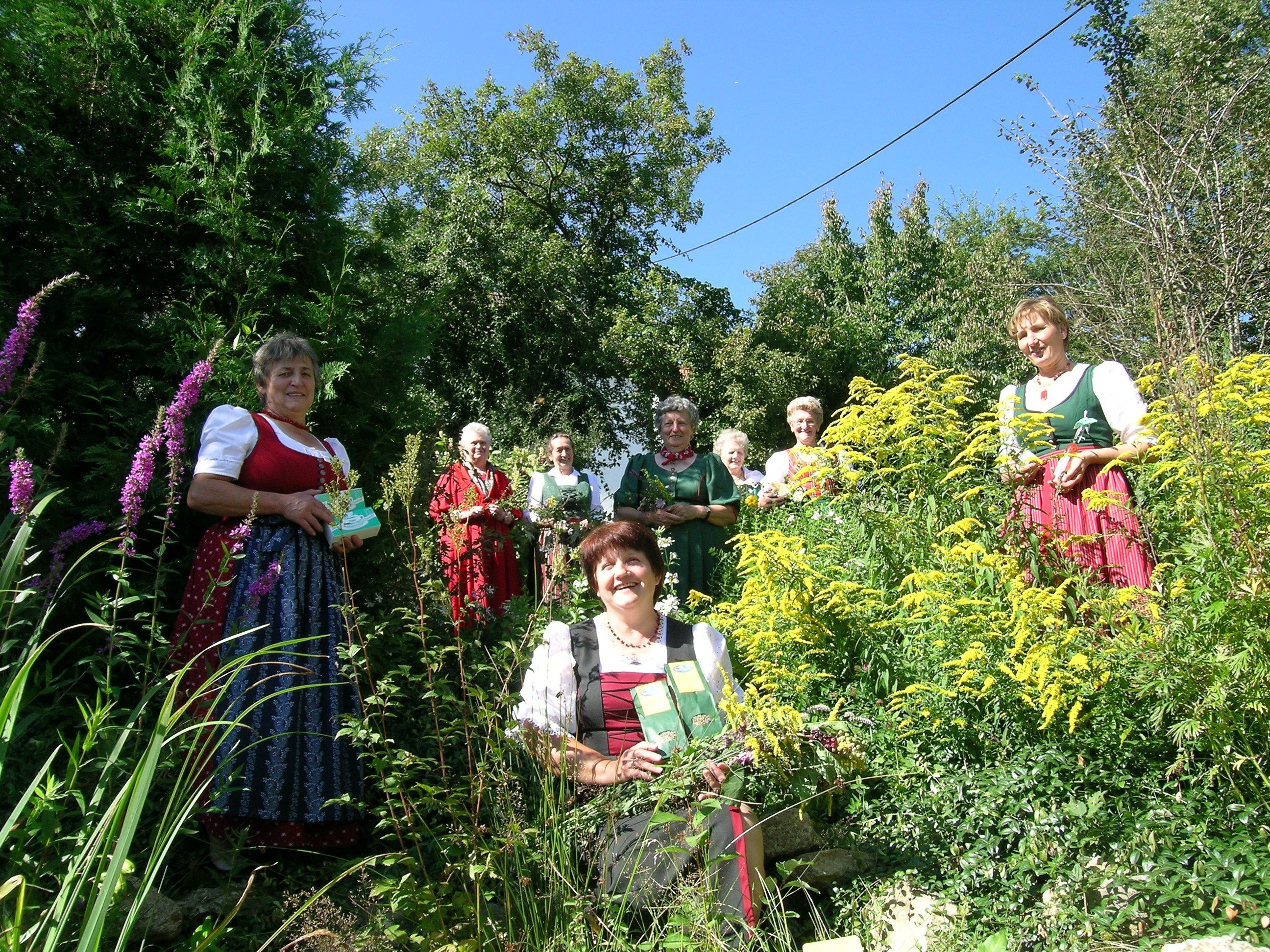 Frauen in traditioneller Kleidung stehen in einem Garten mit Kräutern und Blumen.
