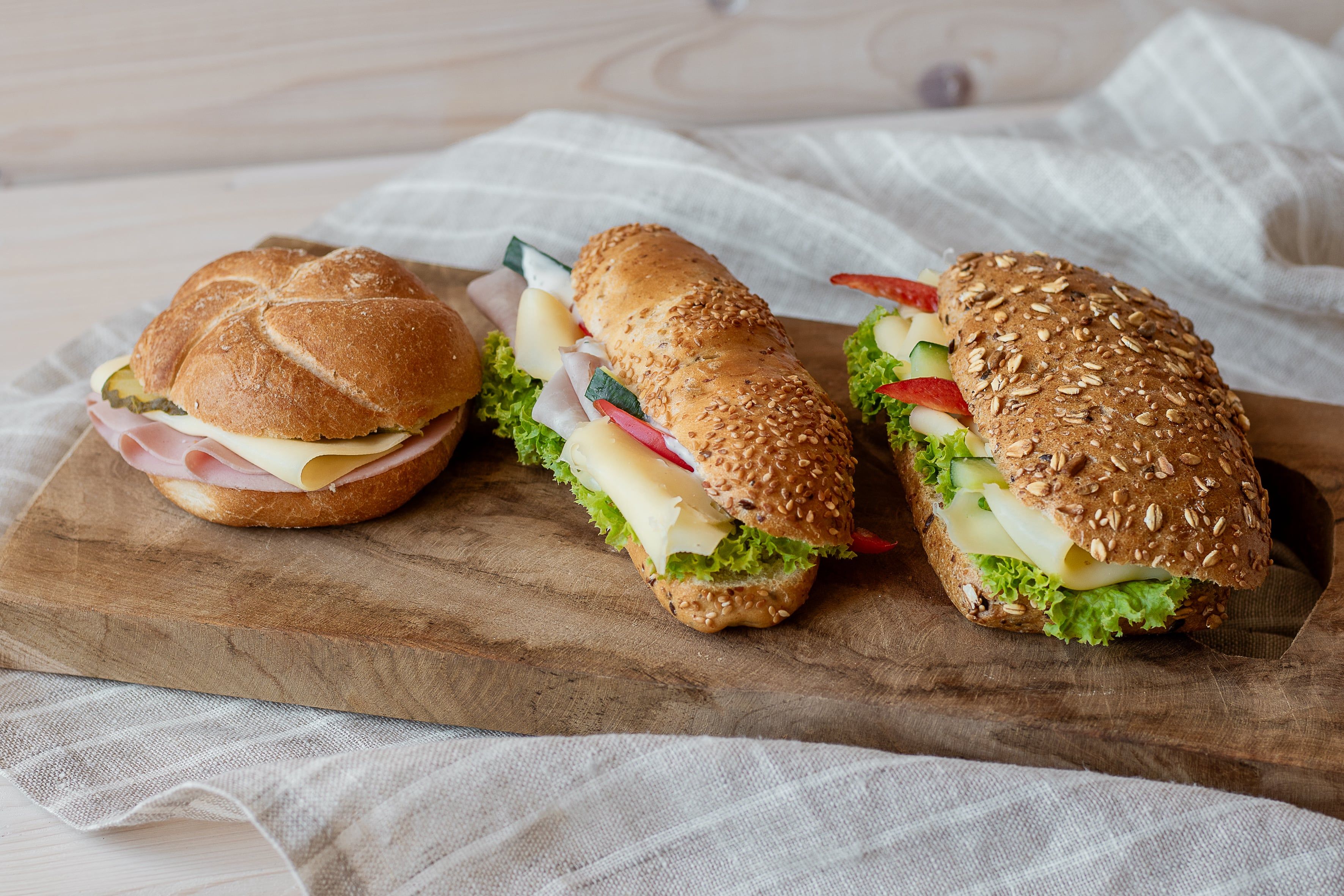 Drei belegte Brötchen auf einem Holzbrett, mit Käse, Schinken und Salat.