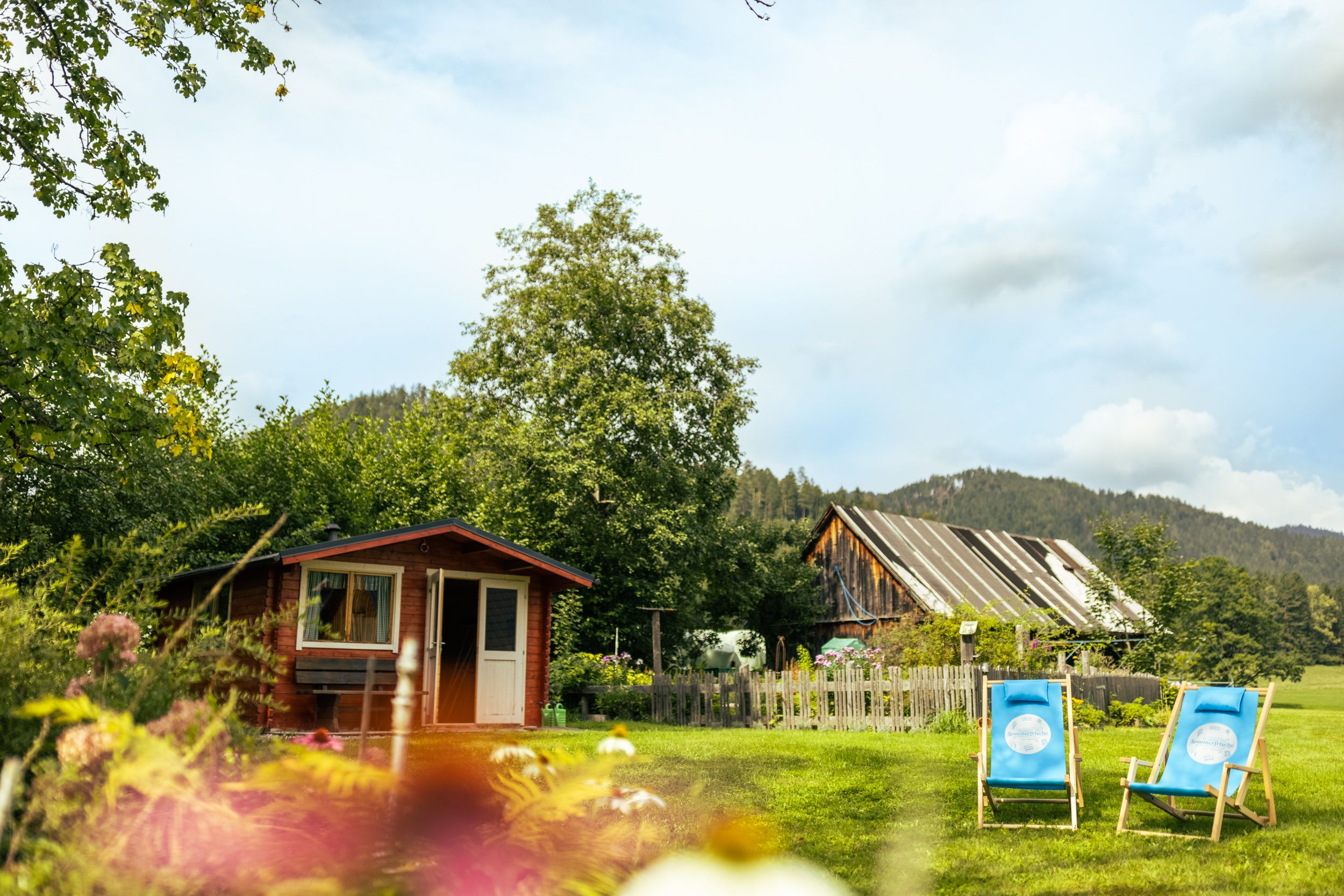 Ein idyllischer Garten mit einem kleinen Holzhaus, zwei blauen Liegestühlen und einem alten Scheunengebäude im Hintergrund.