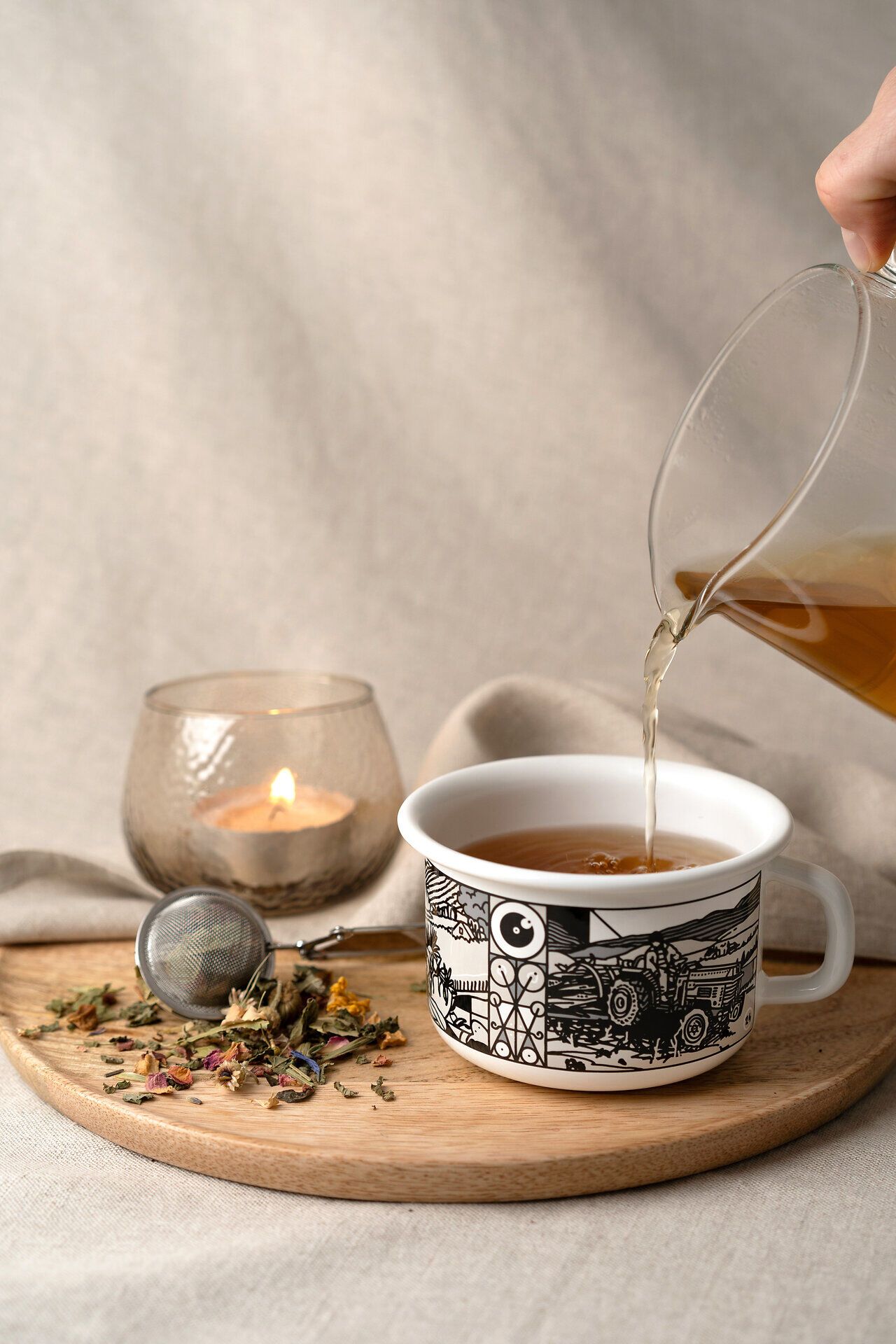 Eine Tasse dampfenden Tees wird liebevoll in eine kunstvoll gestaltete Emailletasse gegossen, während die sanfte Flamme einer Kerze im Hintergrund für eine gemütliche Atmosphäre sorgt. Umgeben von duftenden Kräutern und bunten Blütenblättern lädt dieser Moment zum Entspannen und Genießen ein.