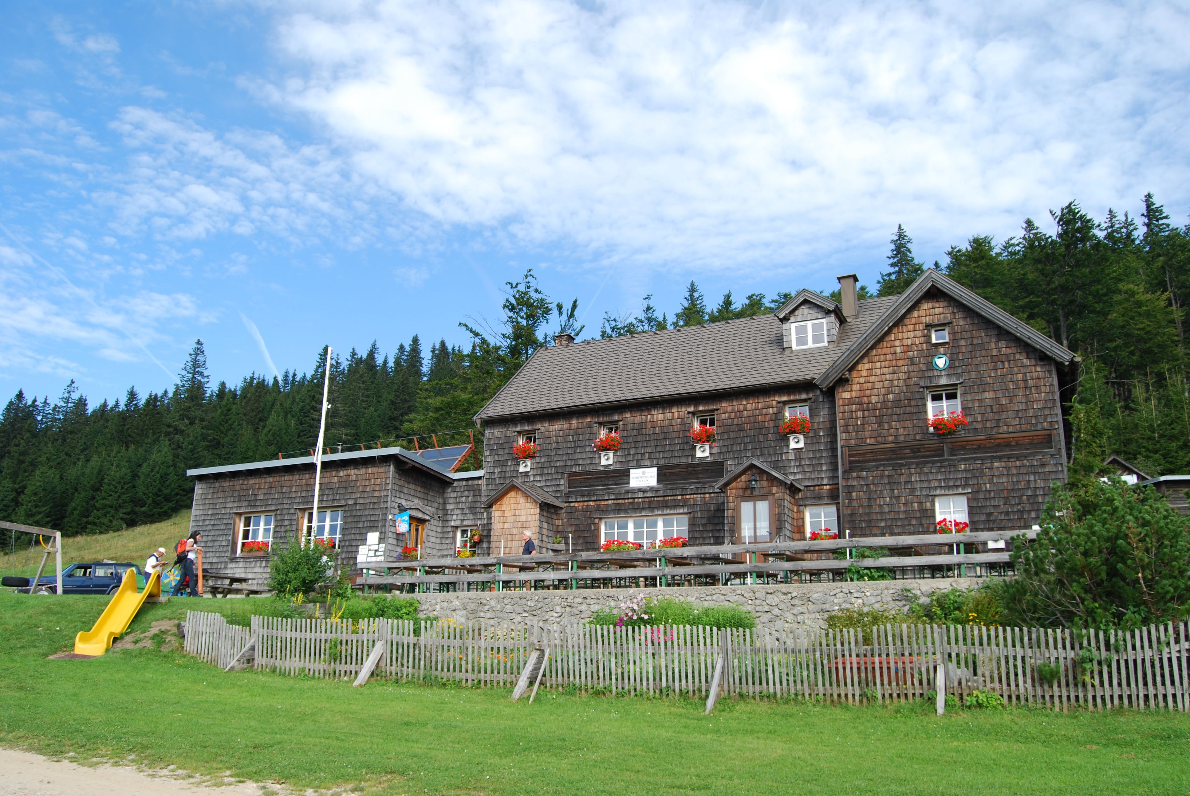 Ein traditionelles Holzhaus mit roten Blumenkästen vor einem Wald und blauem Himmel.