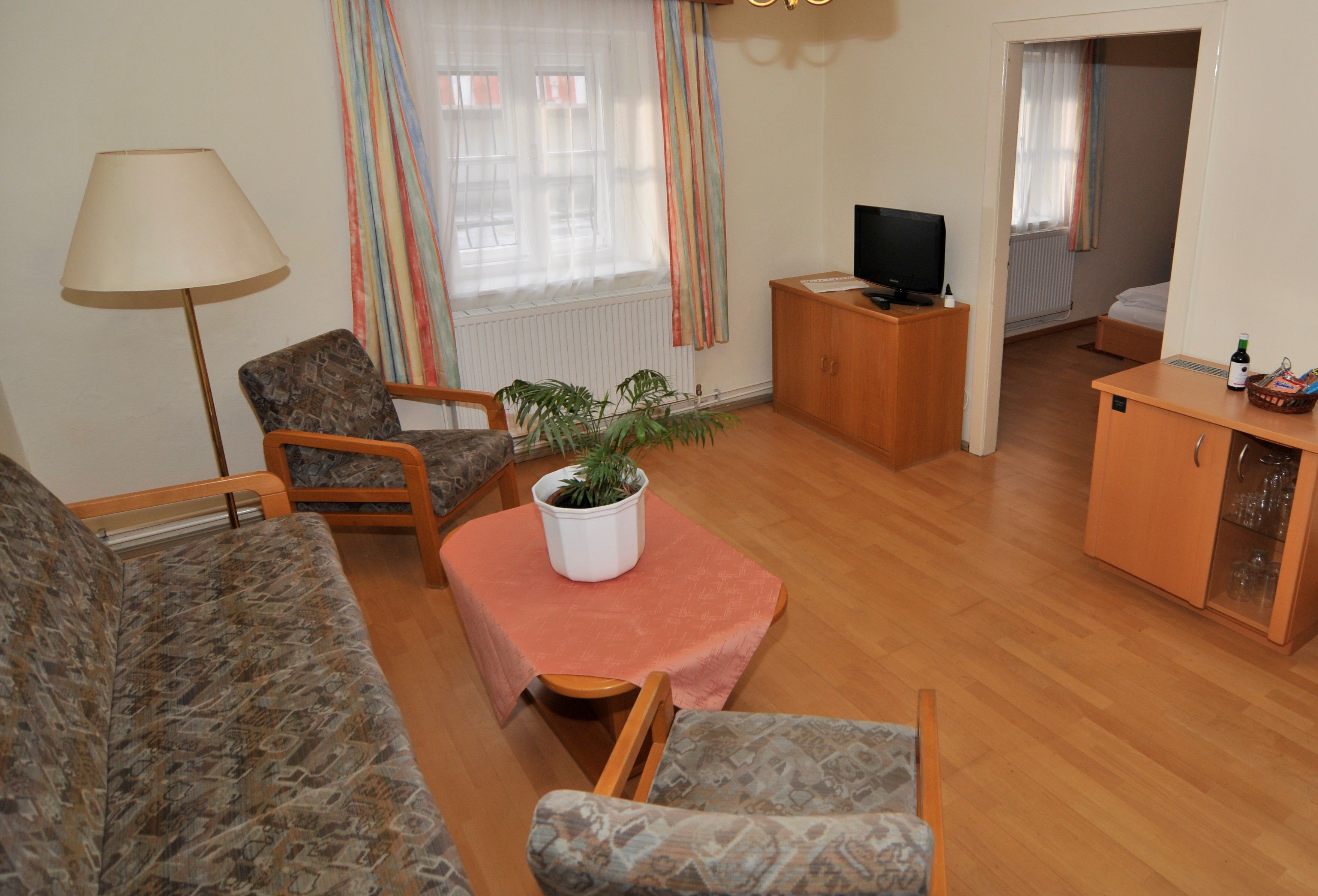 Wohnzimmer mit Sofa, zwei Sesseln, Tisch mit Pflanze, Fernseher und Zugang zu einem Schlafzimmer.