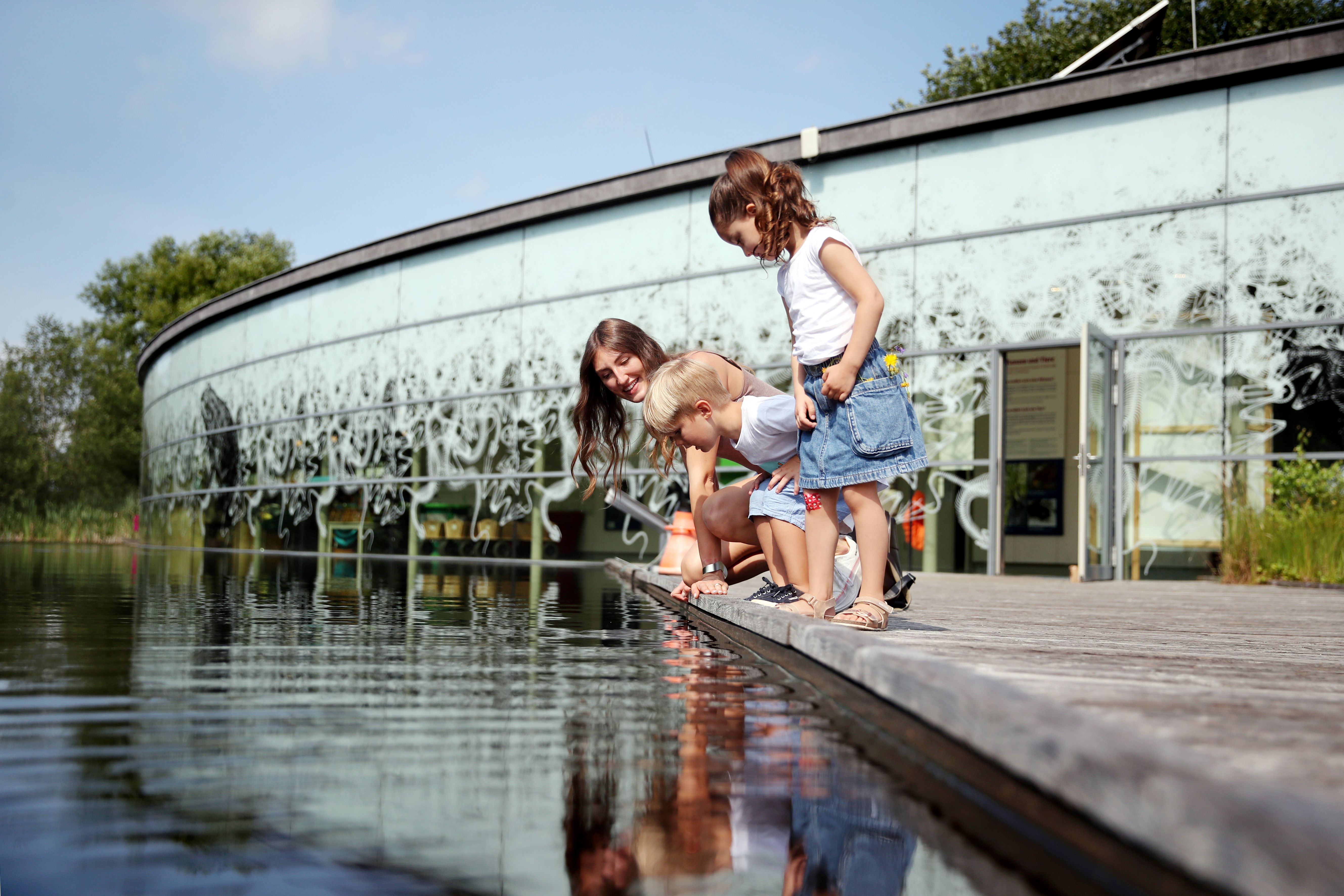 Drei Personen betrachten Wasser an einem Steg vor einem modernen Gebäude.