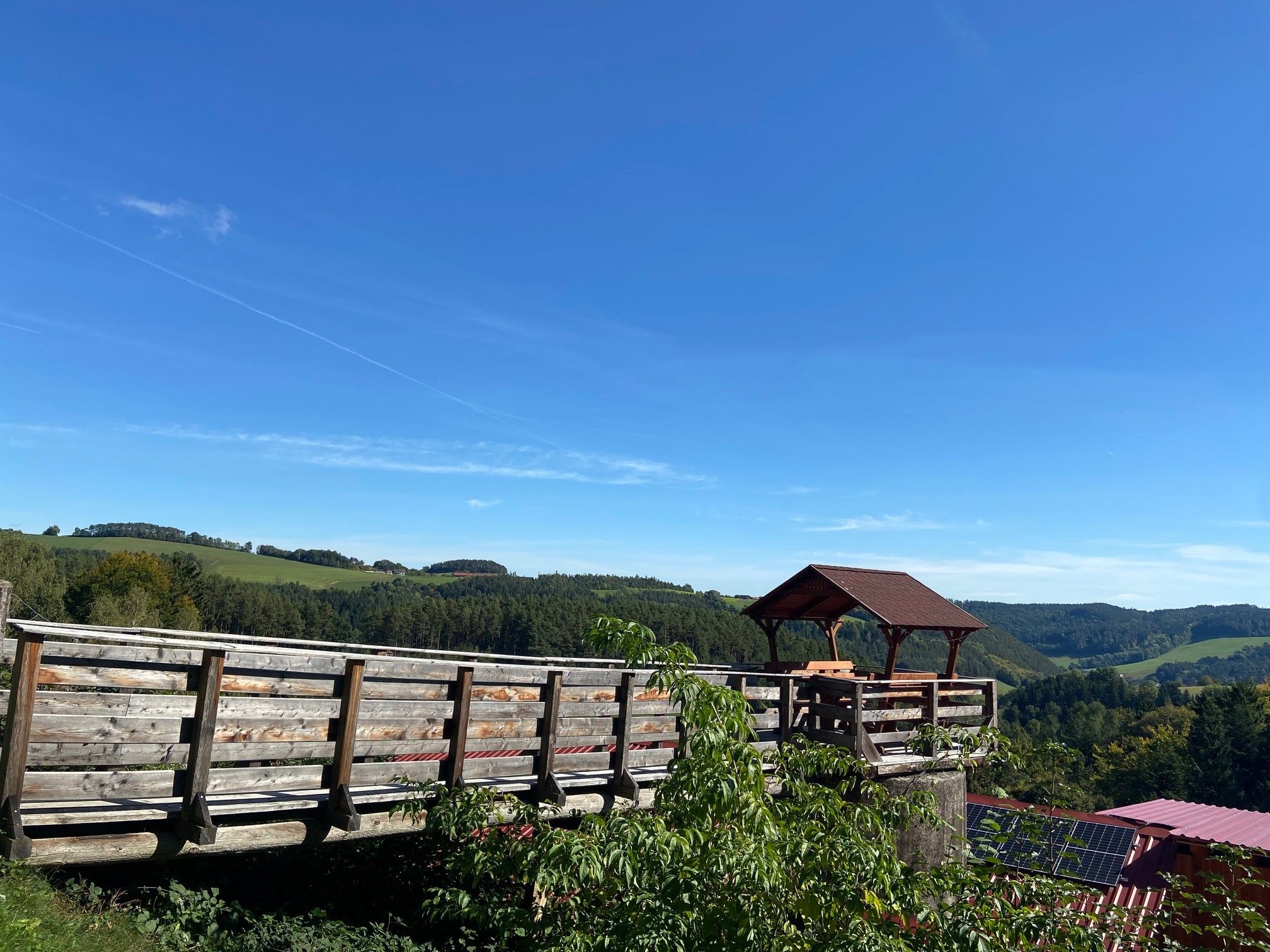 Holzplattform mit Pavillon und Aussicht auf grüne Hügel und blauen Himmel.