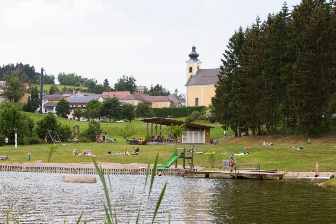 Ein Badeteich mit Liegewiese, Spielplatz und einer Kirche im Hintergrund.