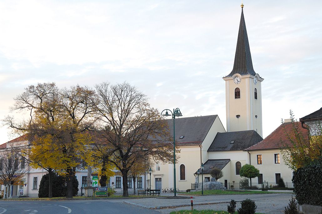 Kirche in Absdorf mit Uhrturm und umliegenden Gebäuden, Bäume im Vordergrund.