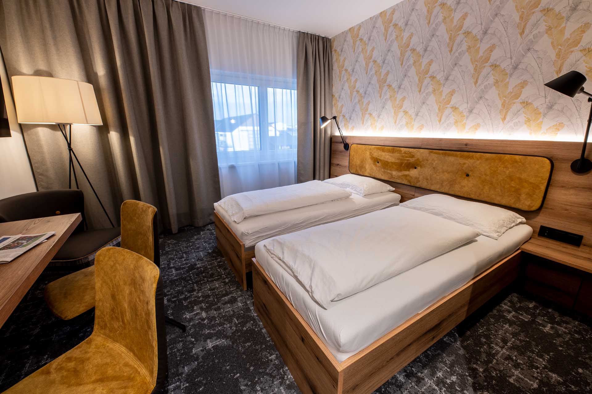 Modernes Gästezimmer mit zwei Einzelbetten, Holzmöbeln, gelben Stühlen und einer Stehlampe. Helle Vorhänge und gemusterte Tapete an der Wand.