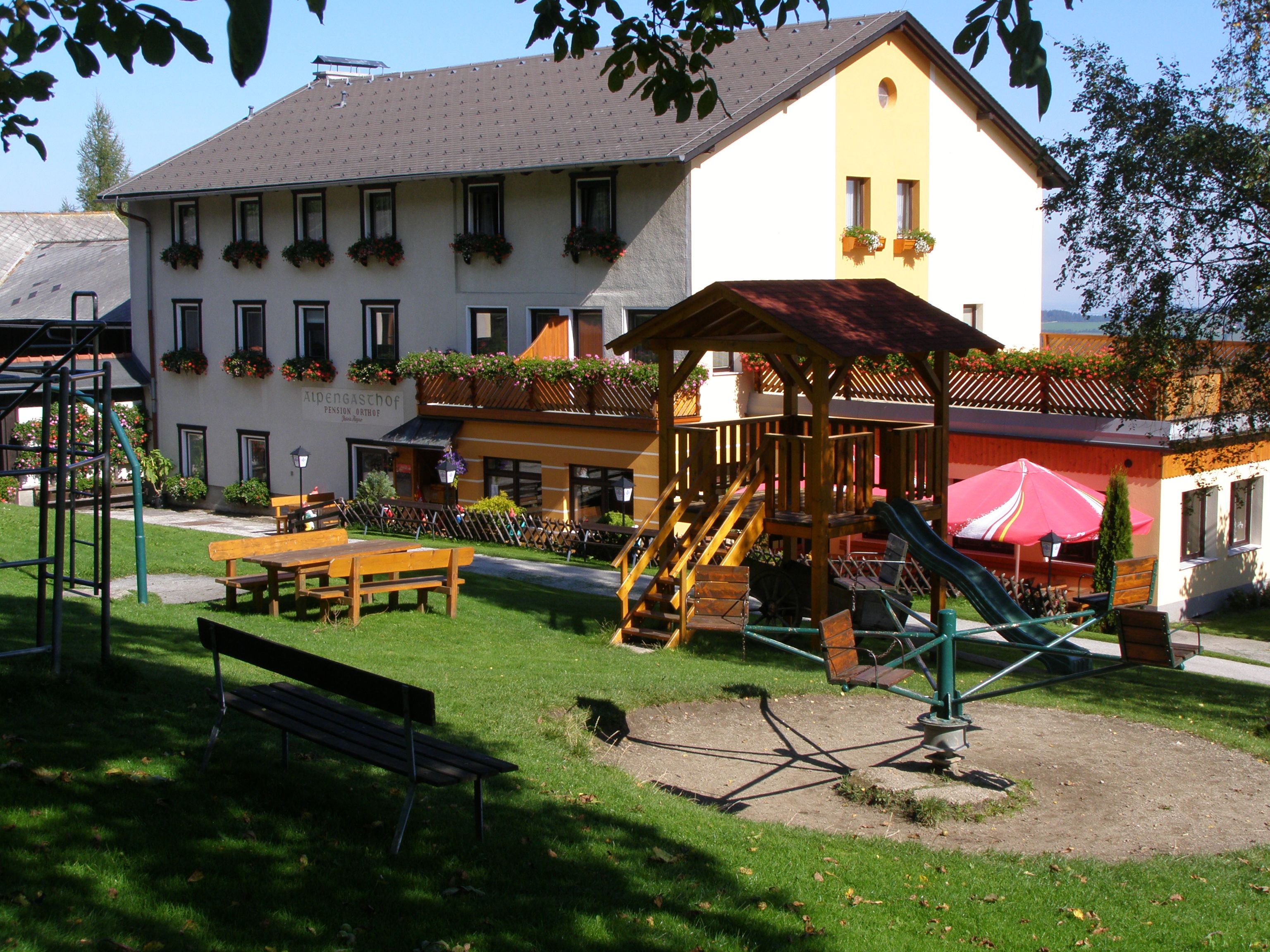 Ein Gasthof mit Spielplatz im Vordergrund, umgeben von grüner Wiese und Bäumen.