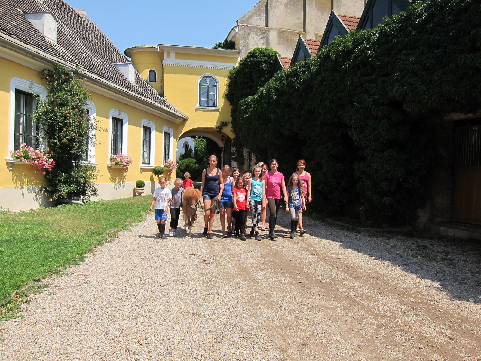 Gruppe von Kindern und Erwachsenen mit Pony vor gelbem Gebäude.