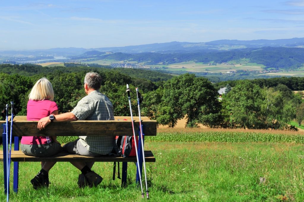 Ein Paar sitzt auf einer Bank mit Wanderstöcken und blickt auf eine hügelige Landschaft.