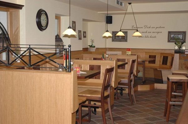 Ein gemütlicher Gastraum mit Holzmöbeln, Tischen und Stühlen. An der Wand hängt eine Uhr und es gibt dekorative Pflanzen.