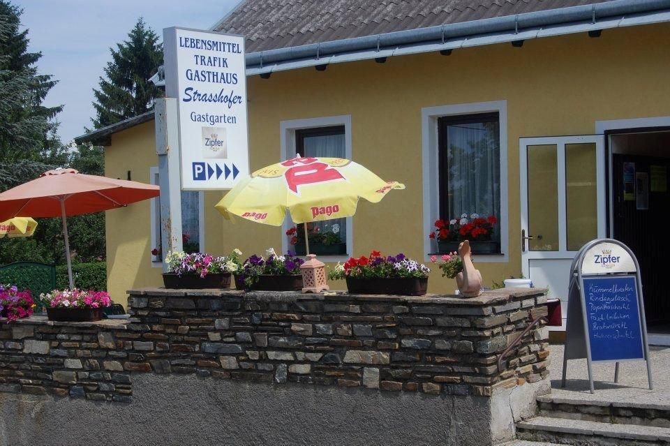 Außenansicht eines gelben Gebäudes mit Schildern für Lebensmittel, Trafik und Gasthaus. Vor dem Haus stehen Sonnenschirme und Blumen.