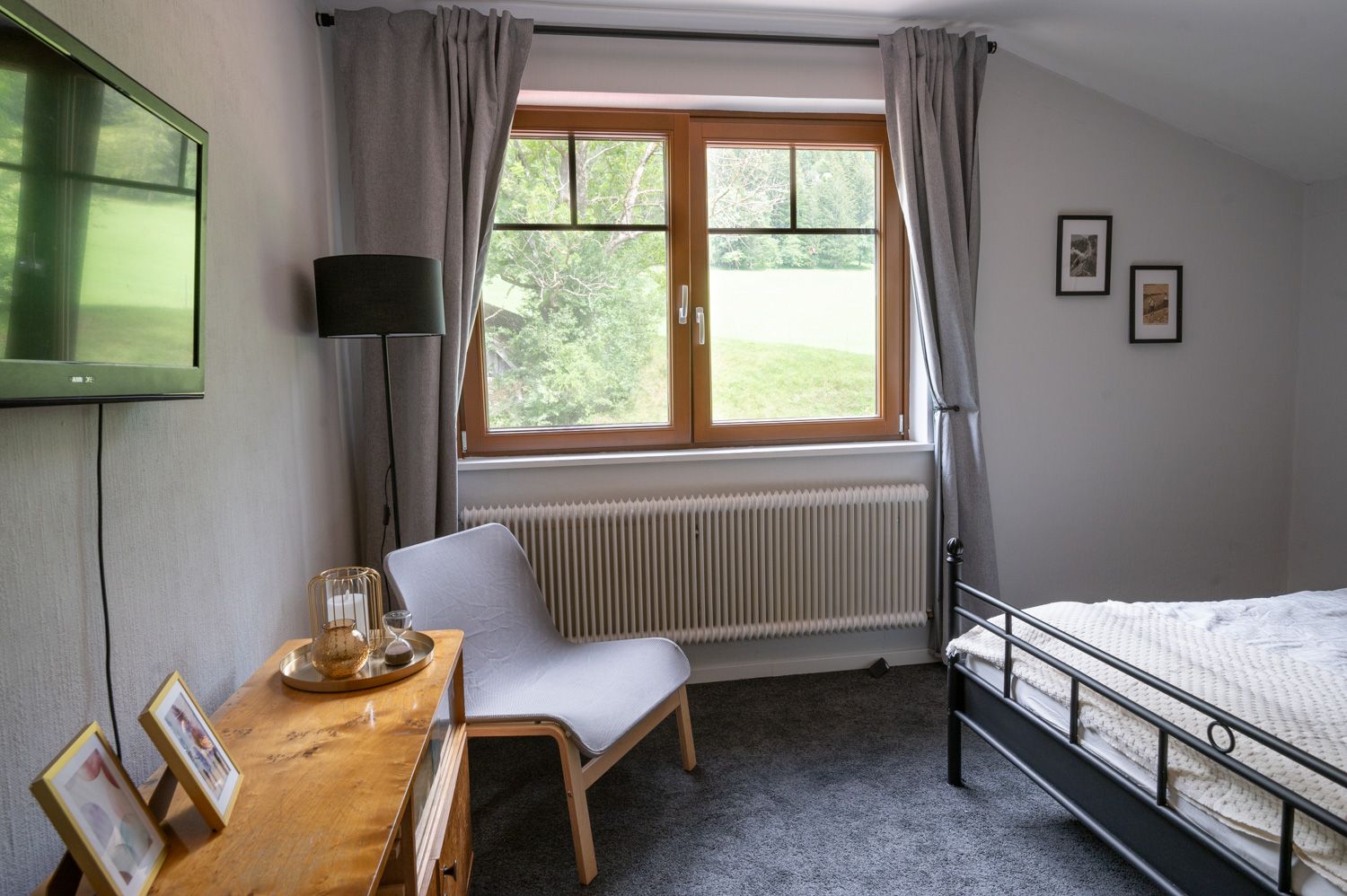 Schlafzimmer mit grauem Teppich, Bett, Sessel, Fernseher und Fenster mit Blick ins Grüne.