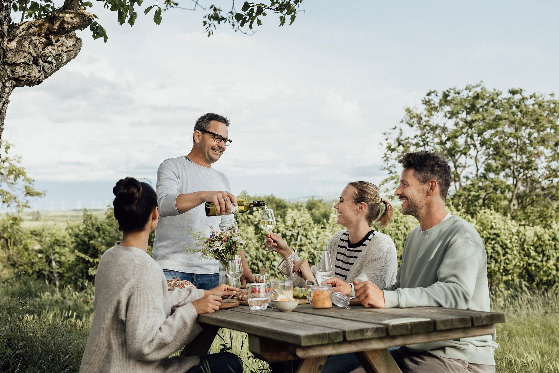Unter einem schattenspendenden Baum versammeln sich Freunde, um eine genussvolle Jause zu teilen. Die frische Luft und die malerische Umgebung laden zum Anstoßen und Verweilen ein, während der Winzer Horst Schuckert seine köstlichen Weine einschenkt. Hier wird der Weinfrühling in vollen Zügen gefeiert.