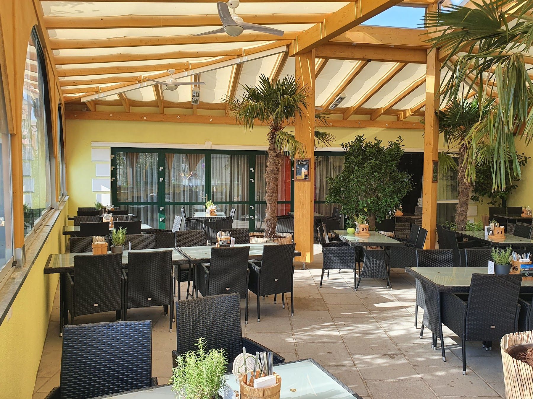 Überdachte Terrasse eines Restaurants mit schwarzen Rattanmöbeln und Pflanzen.