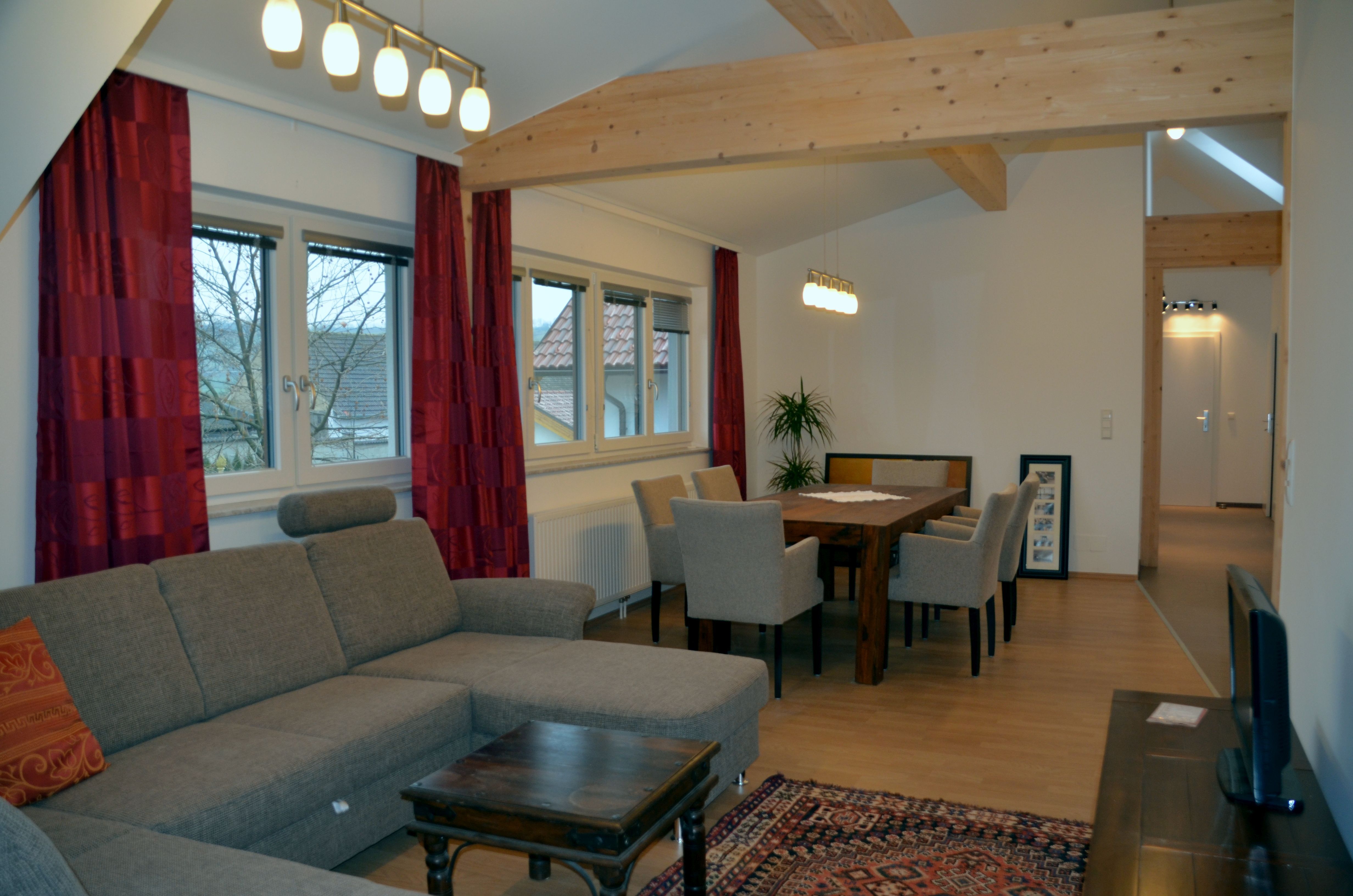Wohnzimmer mit grauem Sofa, Holztisch und Stühlen, roten Vorhängen und Deckenleuchten.