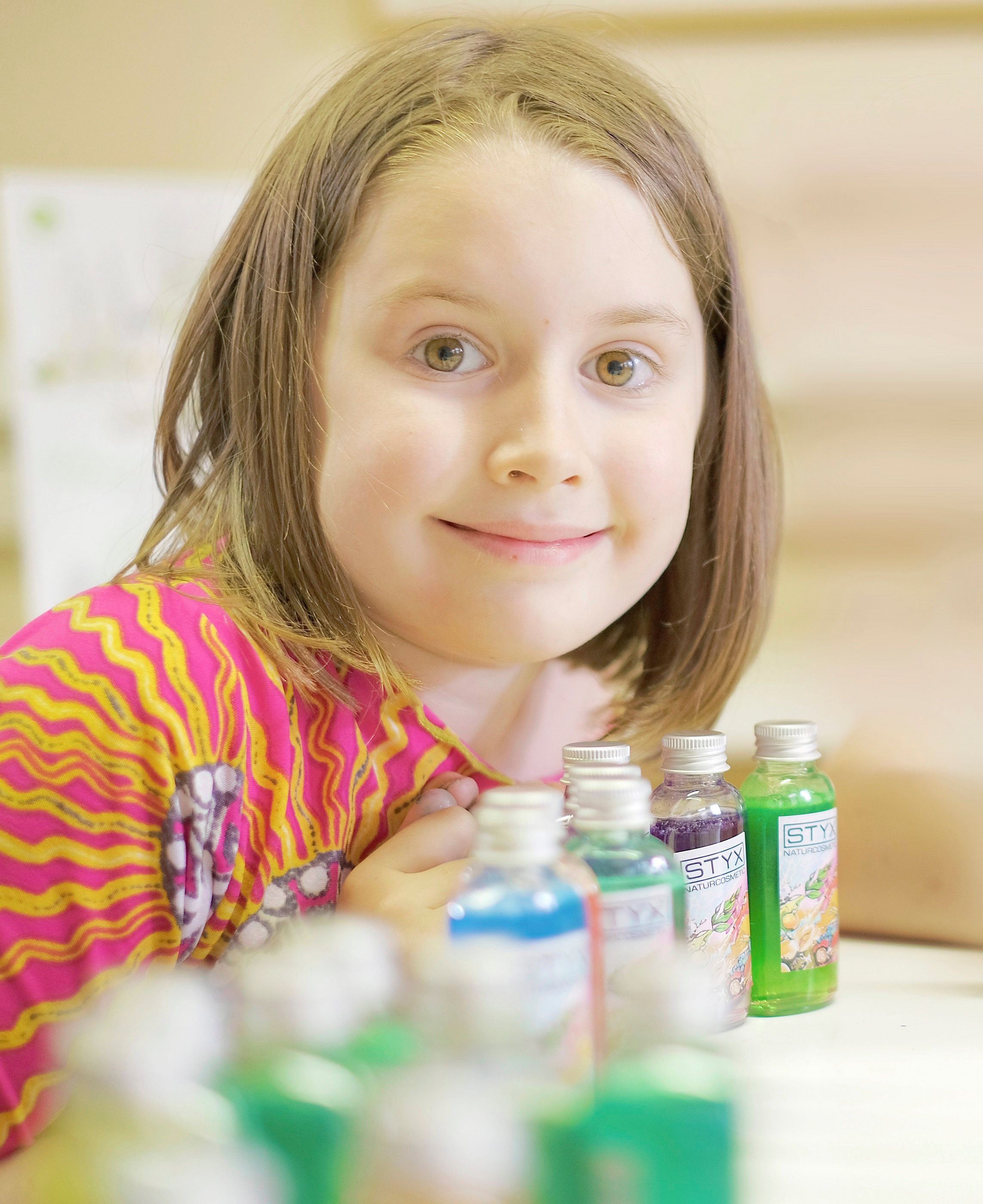 Ein Kind in buntem Oberteil lächelt neben kleinen Flaschen mit Duschgel.