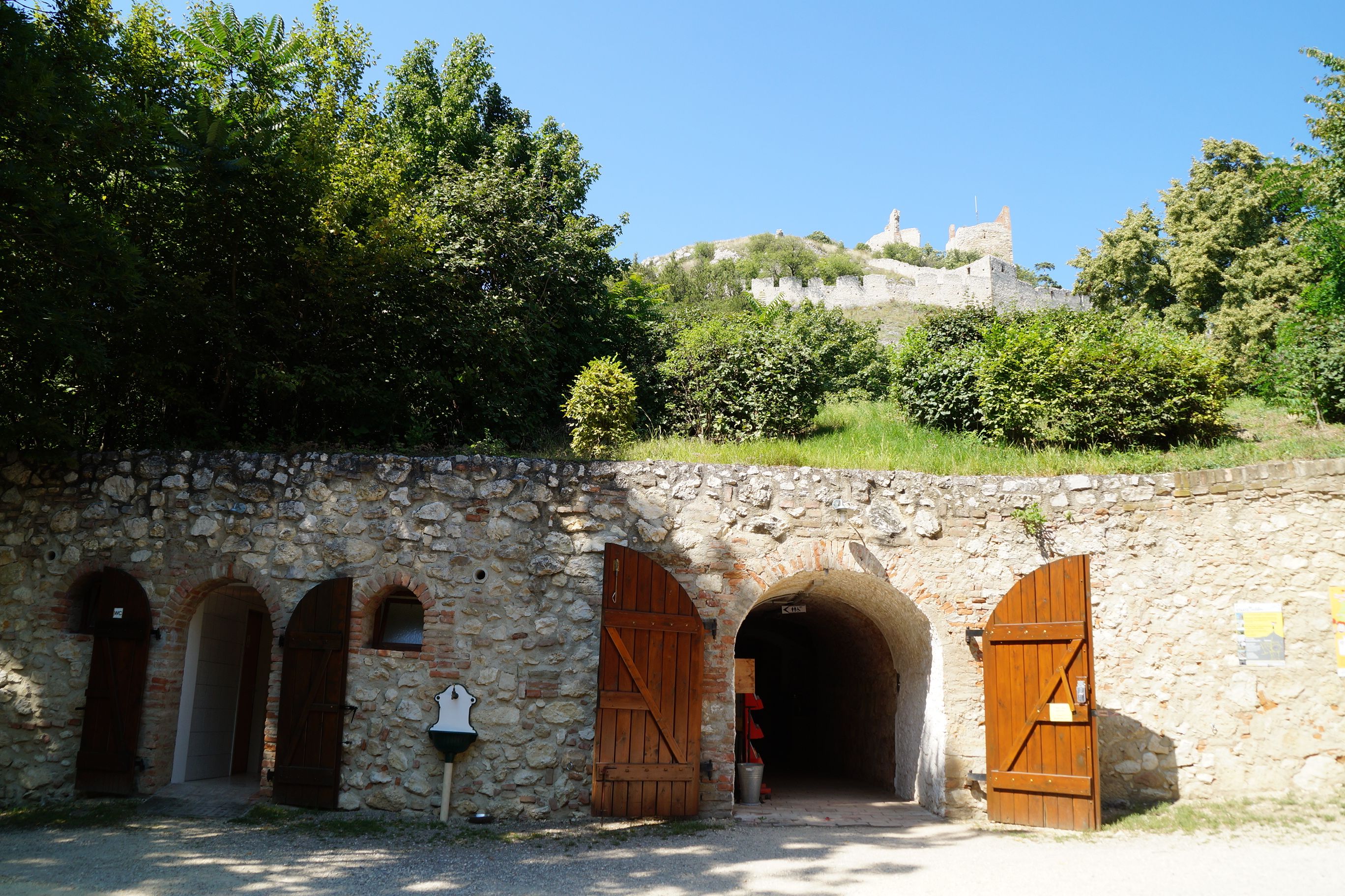 Eingang zu einem Schlosskeller mit Holztüren, darüber eine Burgruine auf einem Hügel.