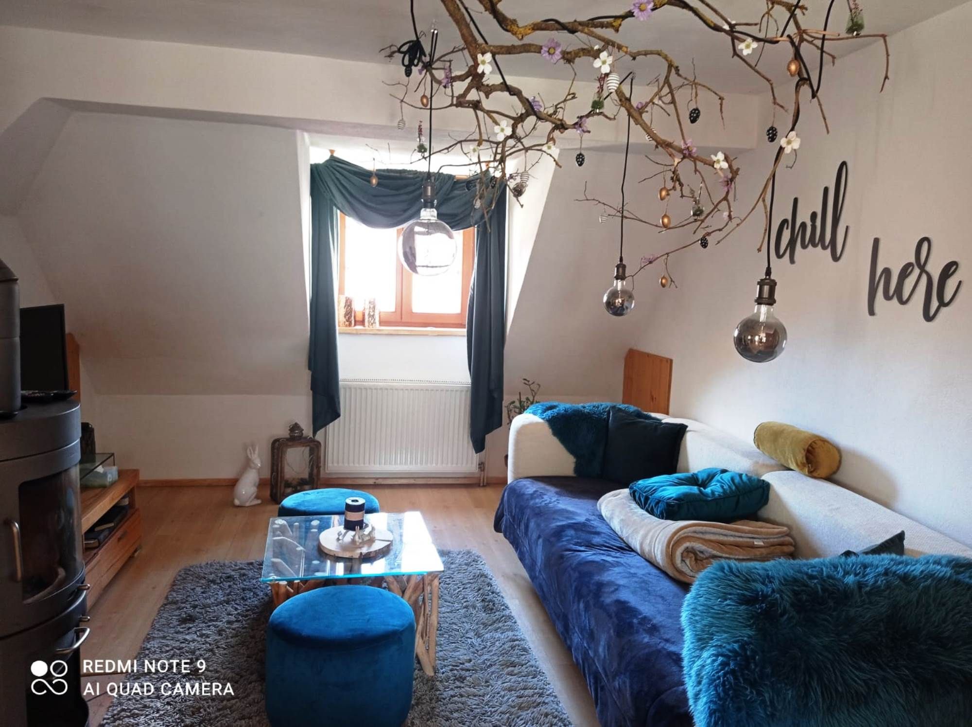 Gemütliches Wohnzimmer mit Sofa, blauem Teppich, Dekozweigen und Schriftzug 'chill here'.