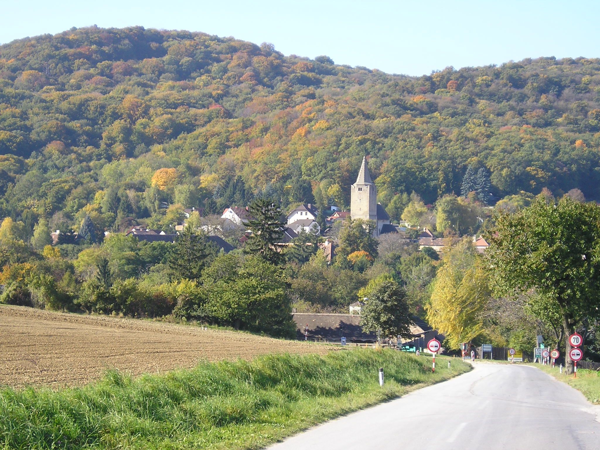 Eine malerische Landschaft mit einer Straße, die zu einem Dorf mit einer Kirche führt, umgeben von bewaldeten Hügeln im Herbst.
