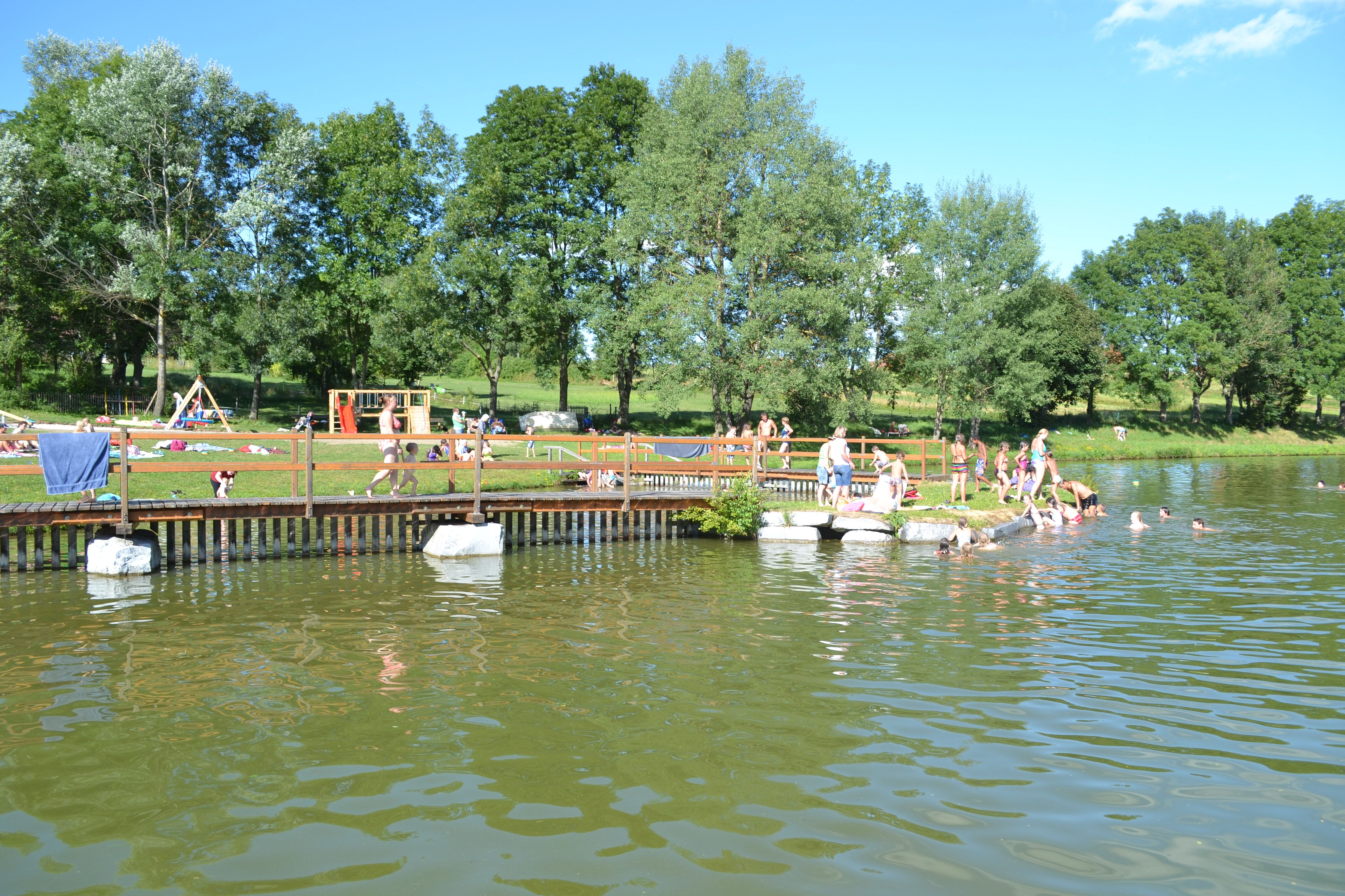 Menschen schwimmen und entspannen an einem Badeteich mit Steg und Spielplatz im Hintergrund.