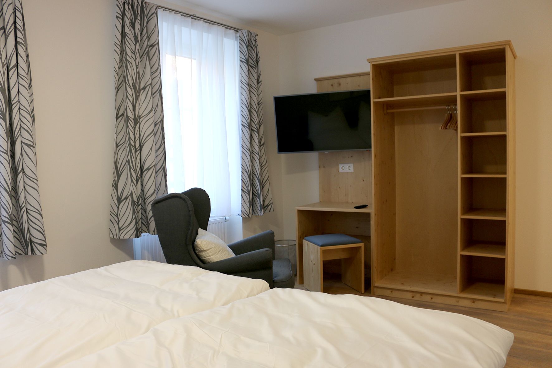 Offener Kleiderschrank mit Fernseher an der Wand. Vor einem Fenster mit einer Gardine steht ein grüner gemütlicher Stuhl. 