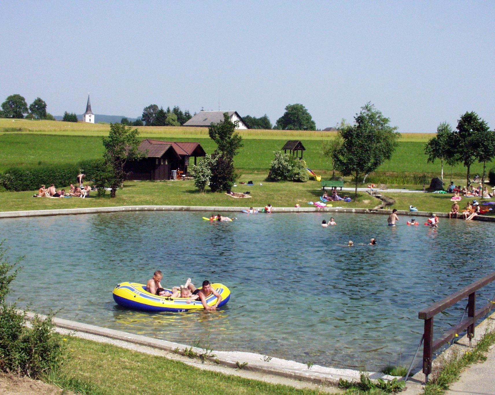 Menschen baden in einem Teich mit umliegender Wiese und Bäumen.