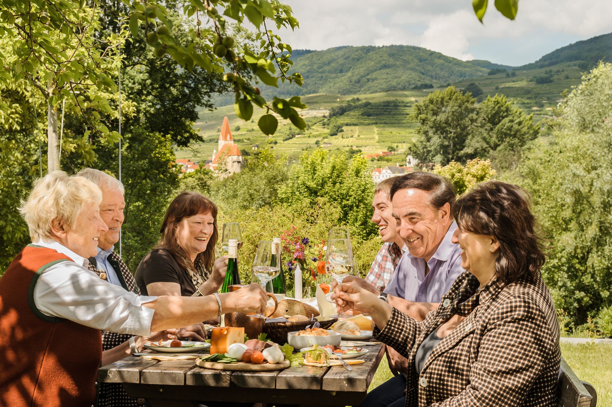 Gruppe von Menschen bei einer Heurigenjause mit Wein, im Hintergrund eine hügelige Landschaft.