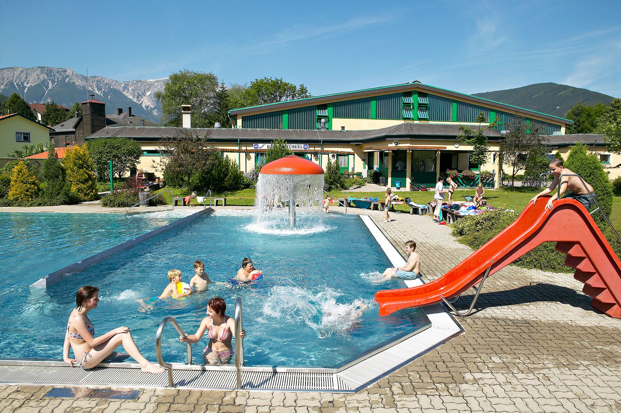 Freibad mit Rutsche und Pilzbrunnen, umgeben von Bergen.