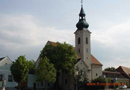 Kirche in Moosbrunn mit grünem Kirchturm und umliegenden Gebäuden.