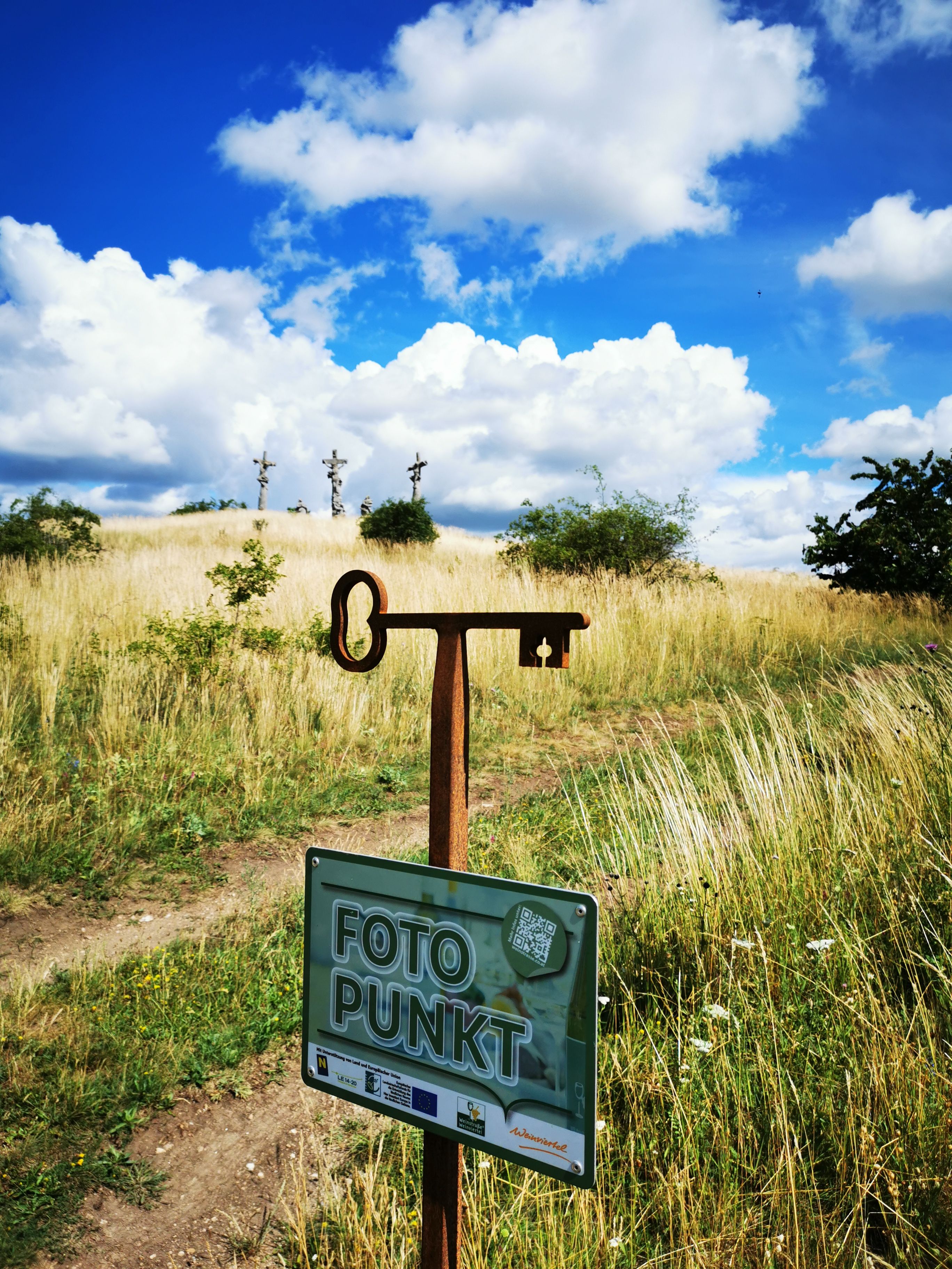 Ein Fotopunkt-Schild vor einer Wiese mit drei Kreuzen im Hintergrund unter blauem Himmel mit Wolken.