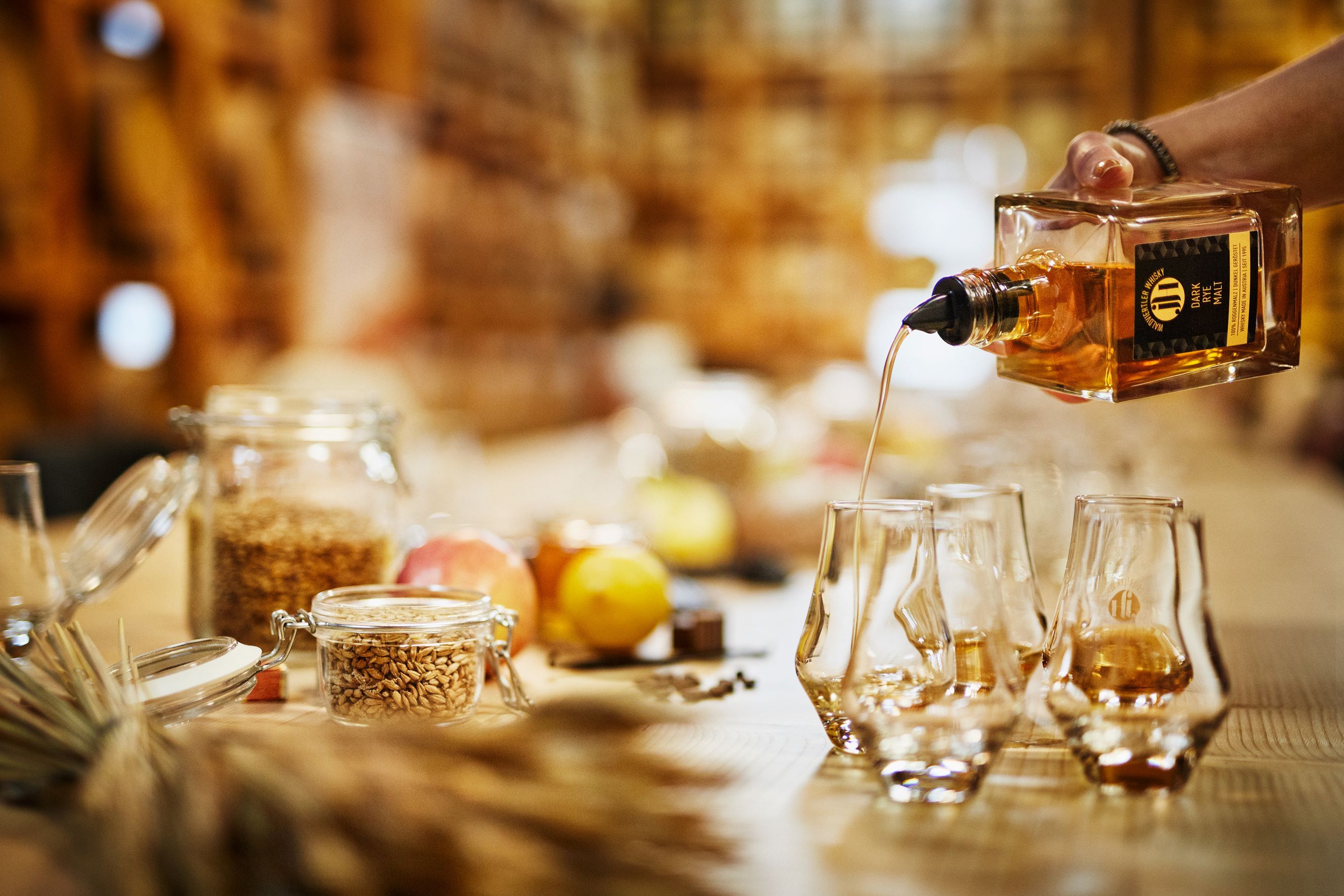 Whisky wird in Gläser gegossen, umgeben von Zutaten wie Getreide und Obst.