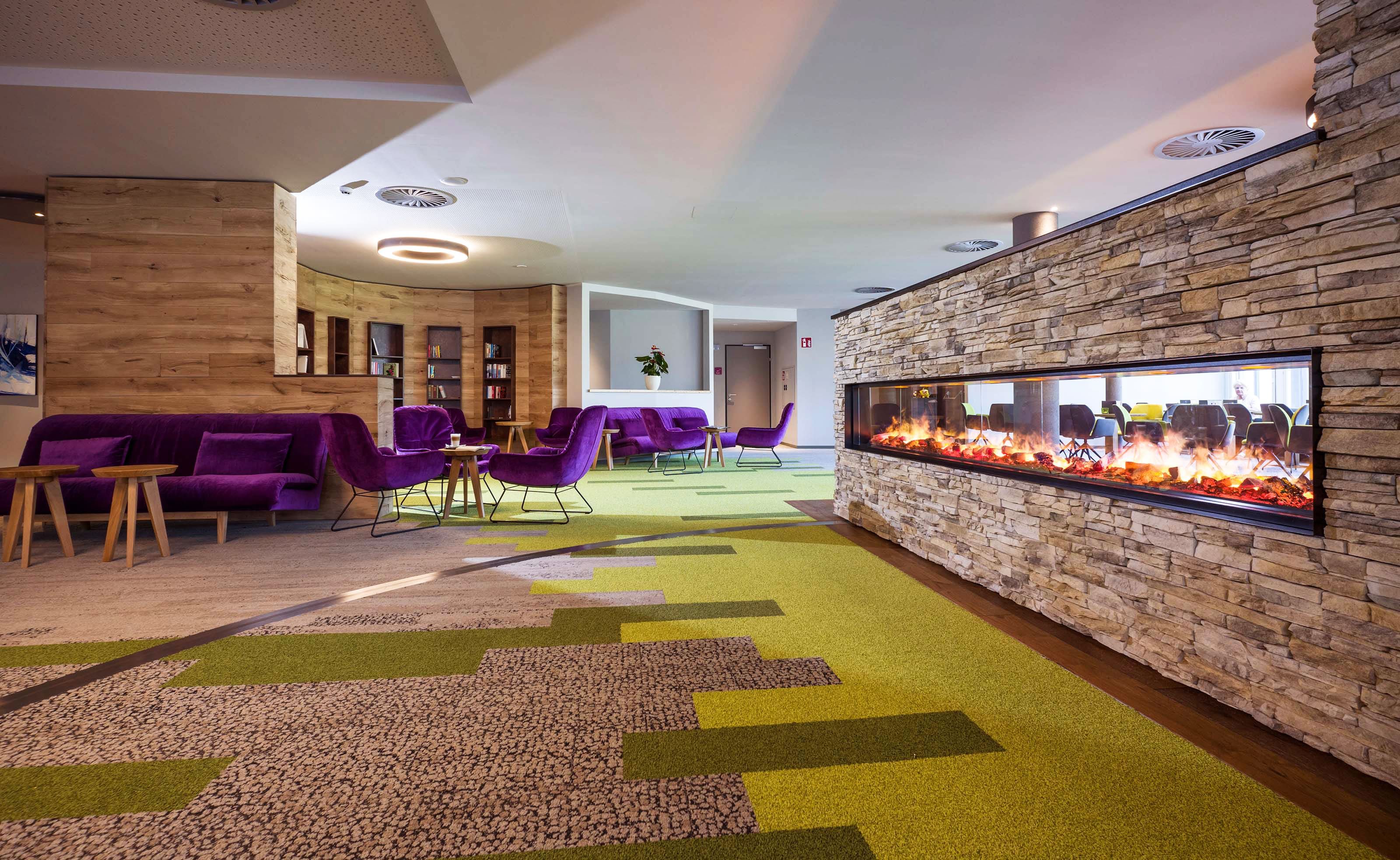 Moderne Hotellobby mit grün-braunem Teppichboden, Kamin, lila Sofas und Stühlen und Holztischen.