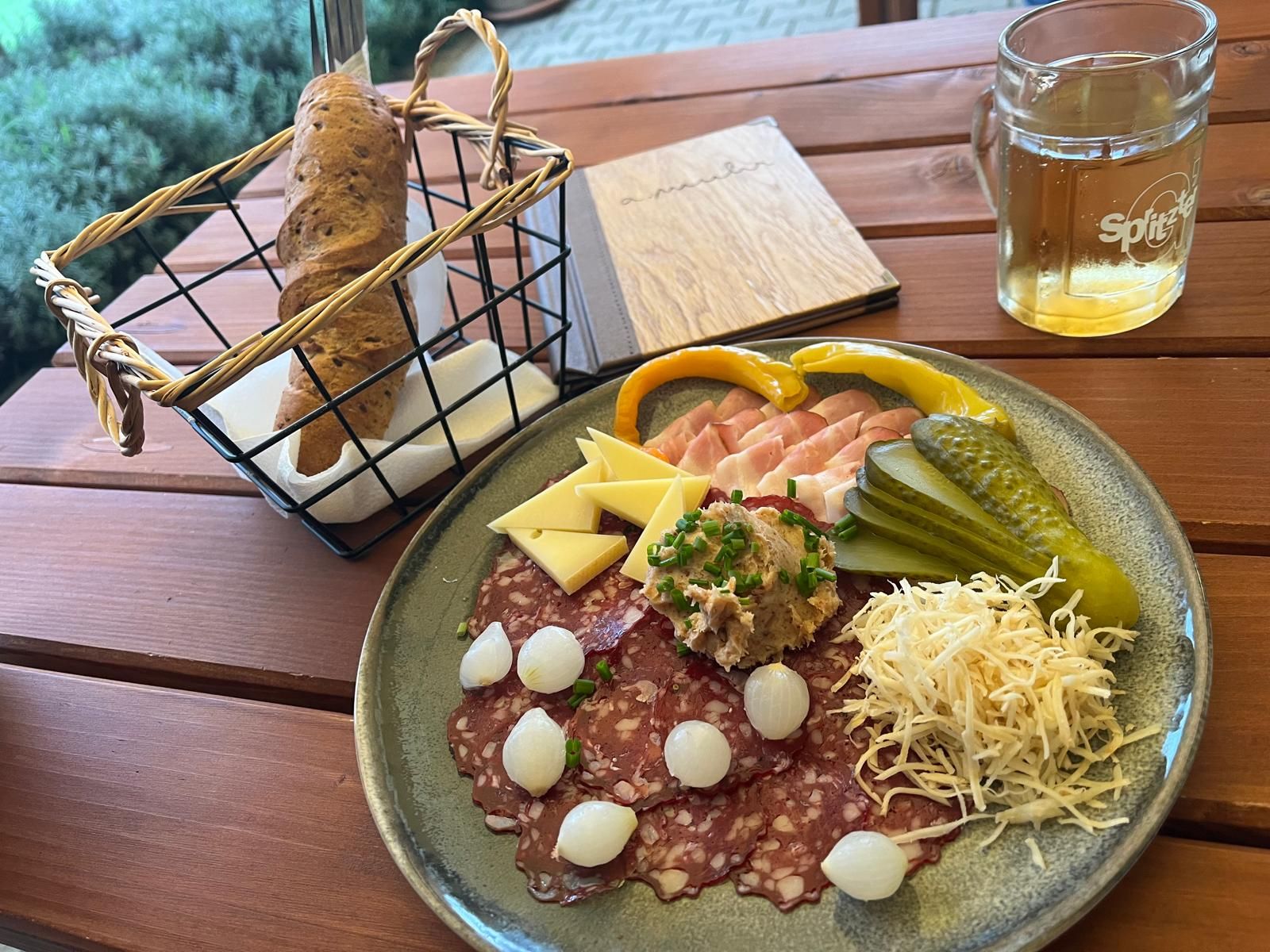 Ein Teller mit Aufschnitt, Käse, Essiggurken und Zwiebeln, daneben ein Brotkorb und ein Getränk auf einem Holztisch.