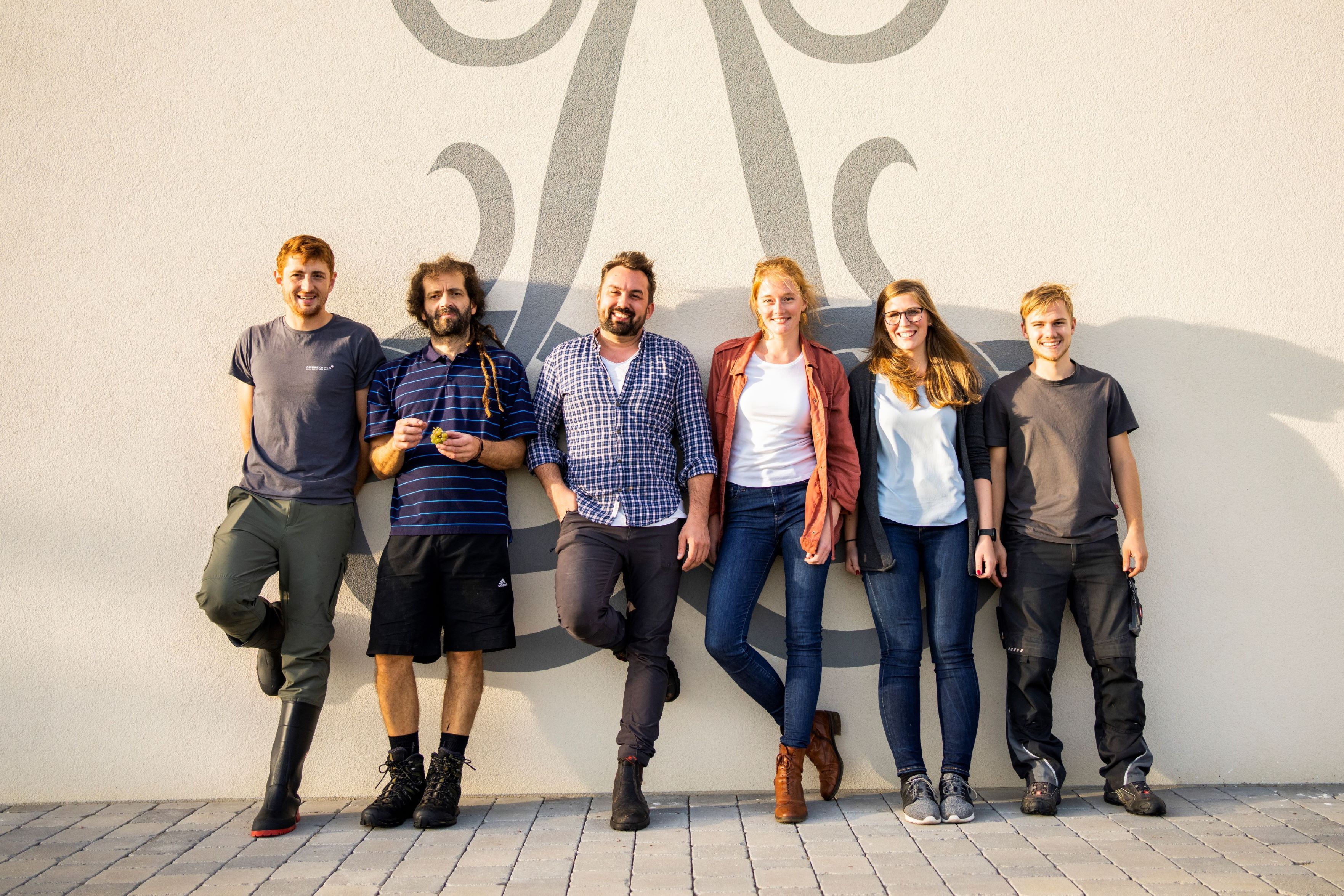 Sechs Personen stehen lächelnd vor einer Wand mit dekorativem Muster.
