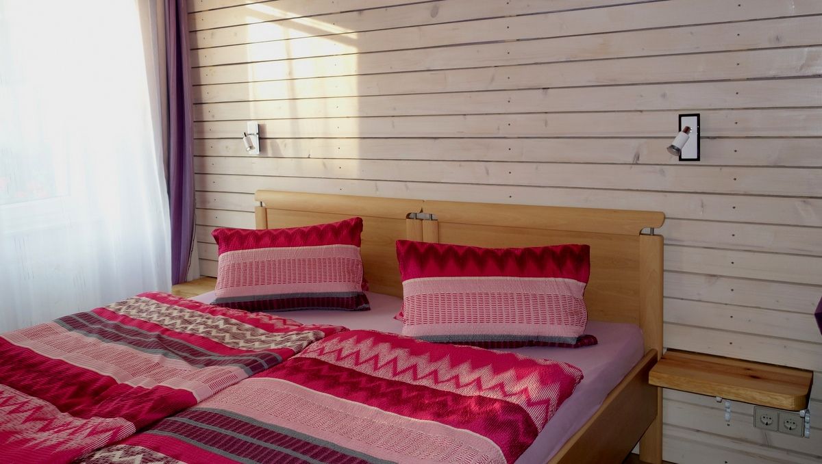 Ein gemütliches Schlafzimmer mit einem Doppelbett, das mit rot-gemusterten Bettdecken und Kissen bezogen ist. Die Wand ist aus hellem Holz.