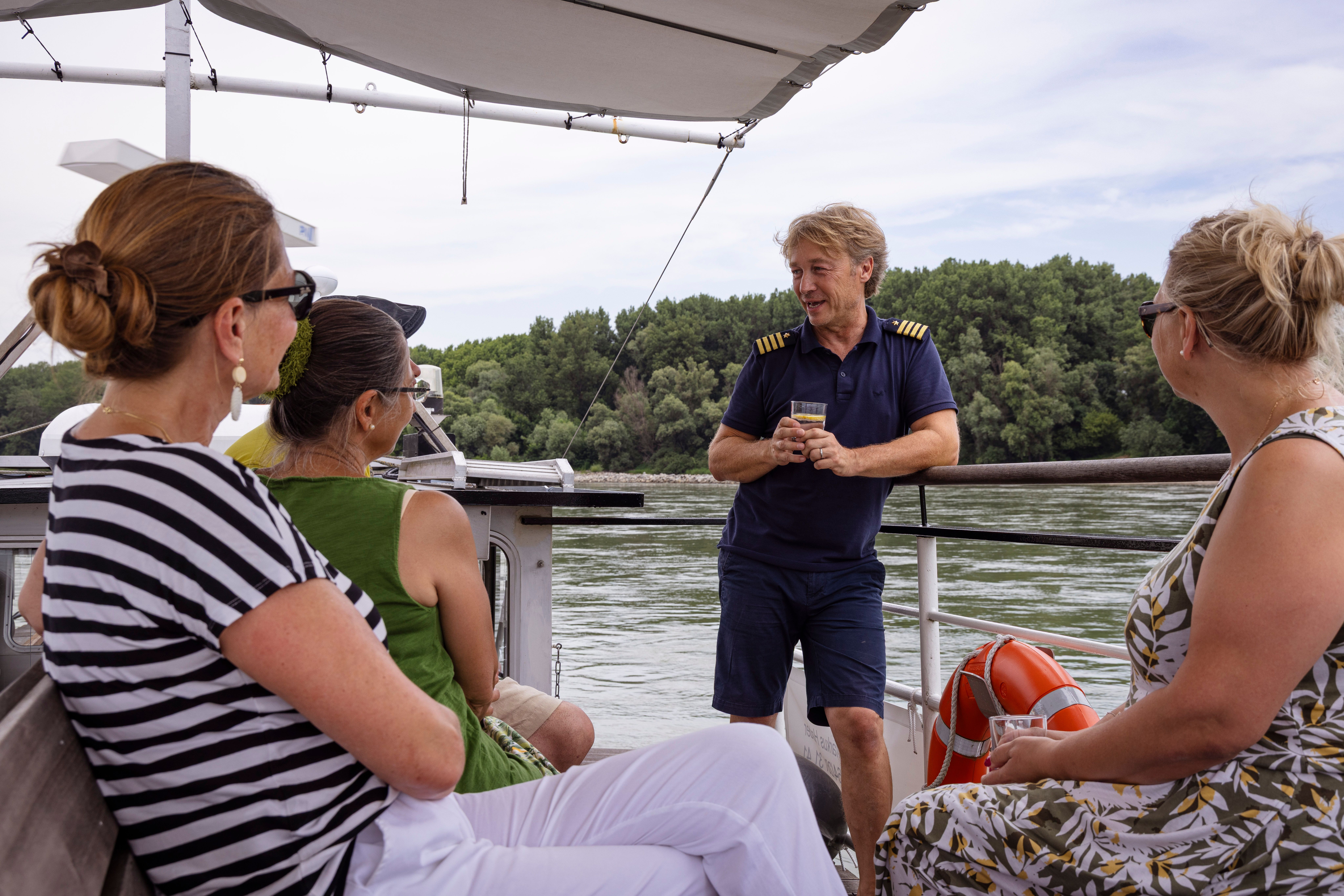 Personen auf einem Boot auf der Donau, ein Mann in Uniform (Kapitän) spricht mit drei Frauen.