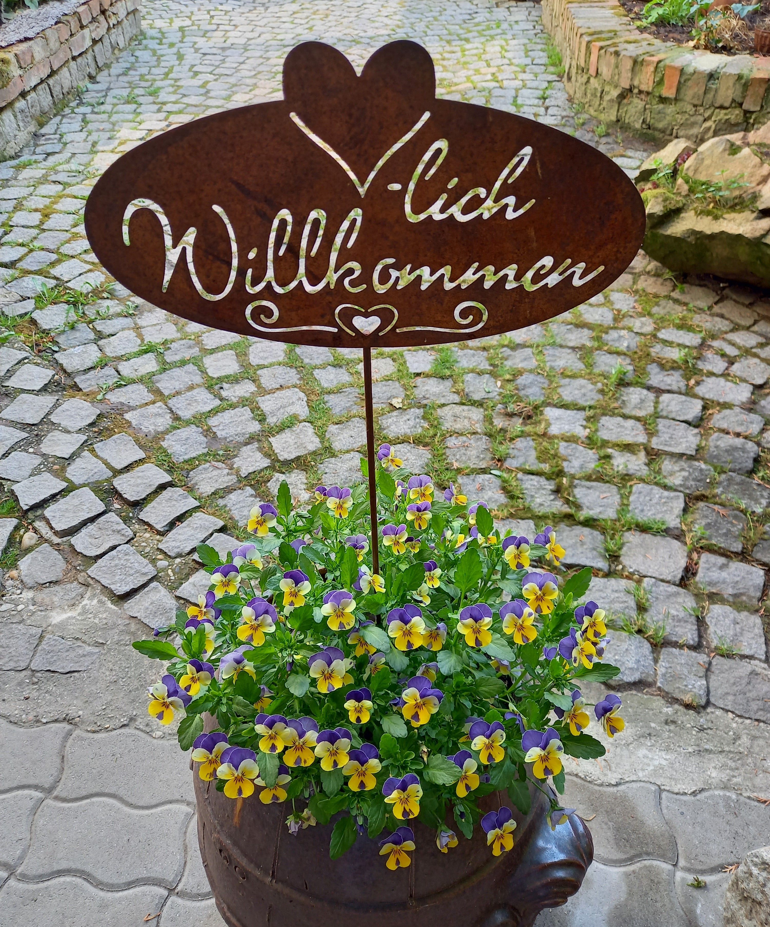 Metallschild mit 'Herzlich Willkommen' über einem Topf mit gelb-violetten Blumen auf einem gepflasterten Weg.