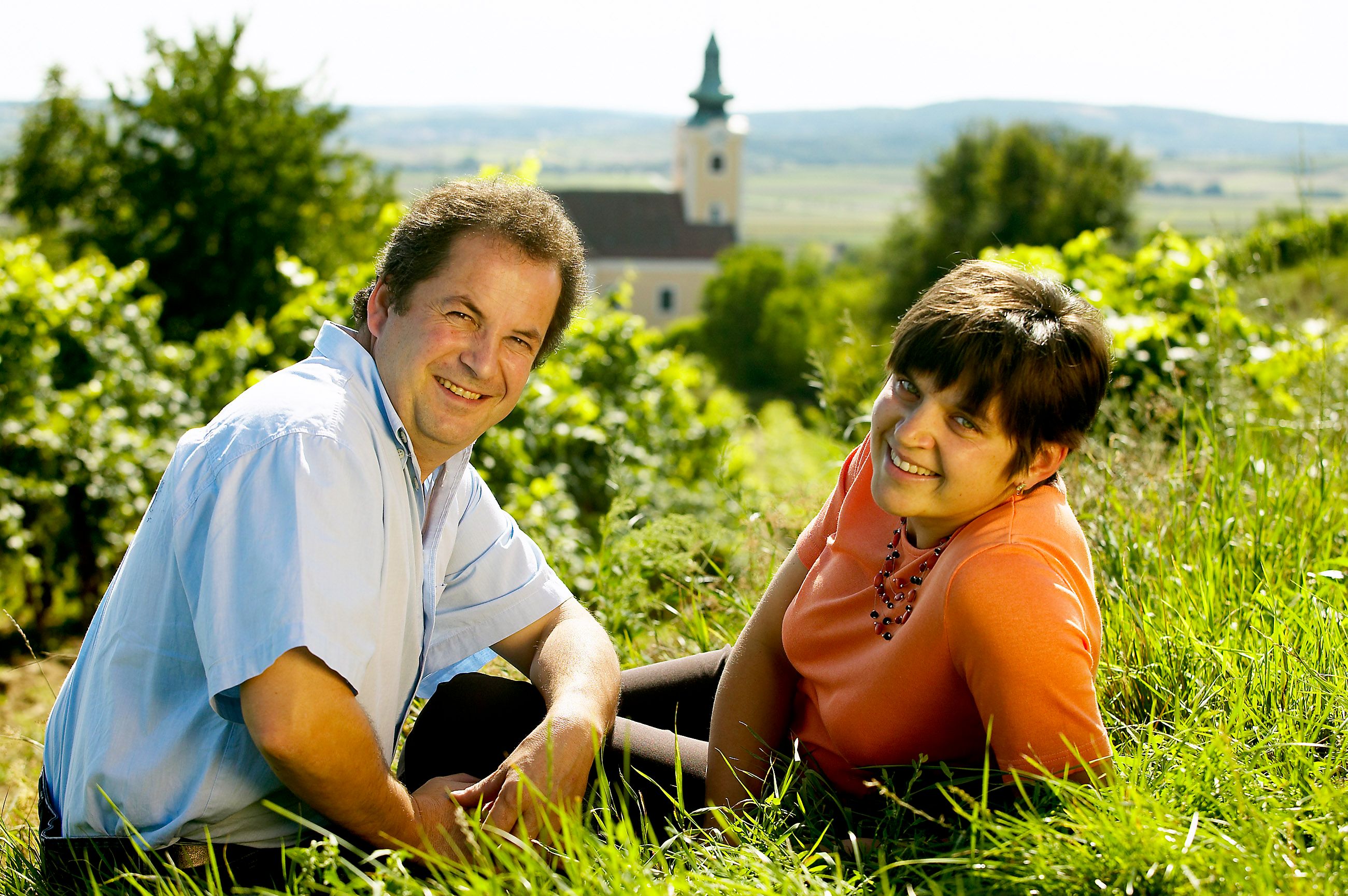 Ein Mann und eine Frau sitzen lächelnd im Gras vor einer Kirche in einer ländlichen Landschaft.