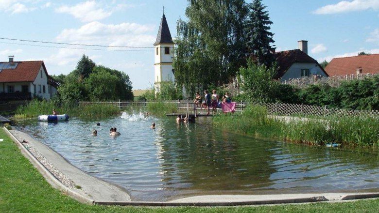 Ein natürlicher Badeteich mit Menschen, die schwimmen. Im Hintergrund sind ein Kirchturm und Häuser zu sehen.