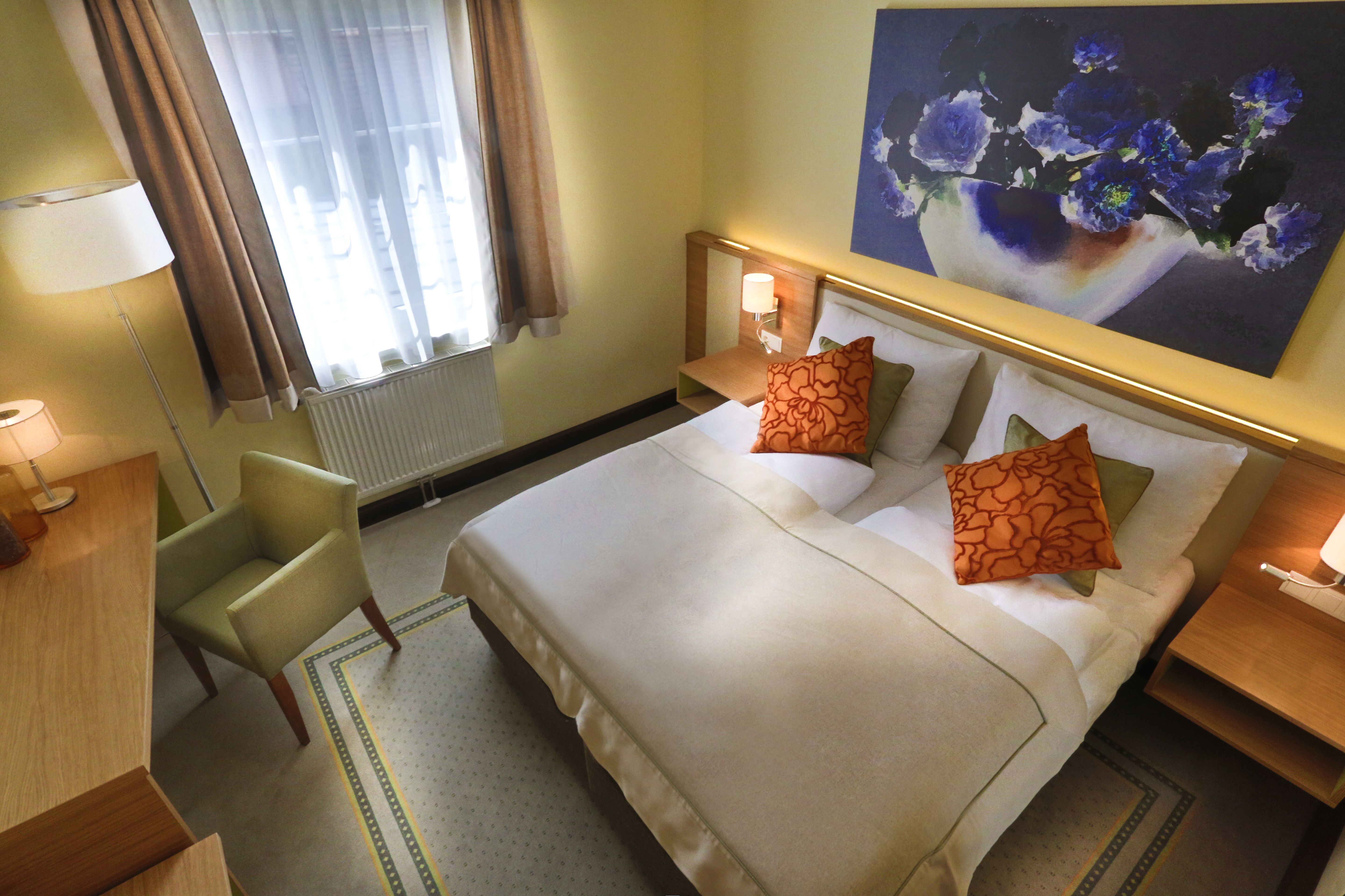 Gemütliches Hotelzimmer mit Doppelbett, zwei orangefarbenen Kissen, einem Schreibtisch, einem Sessel und einem großen Blumenbild an der Wand.