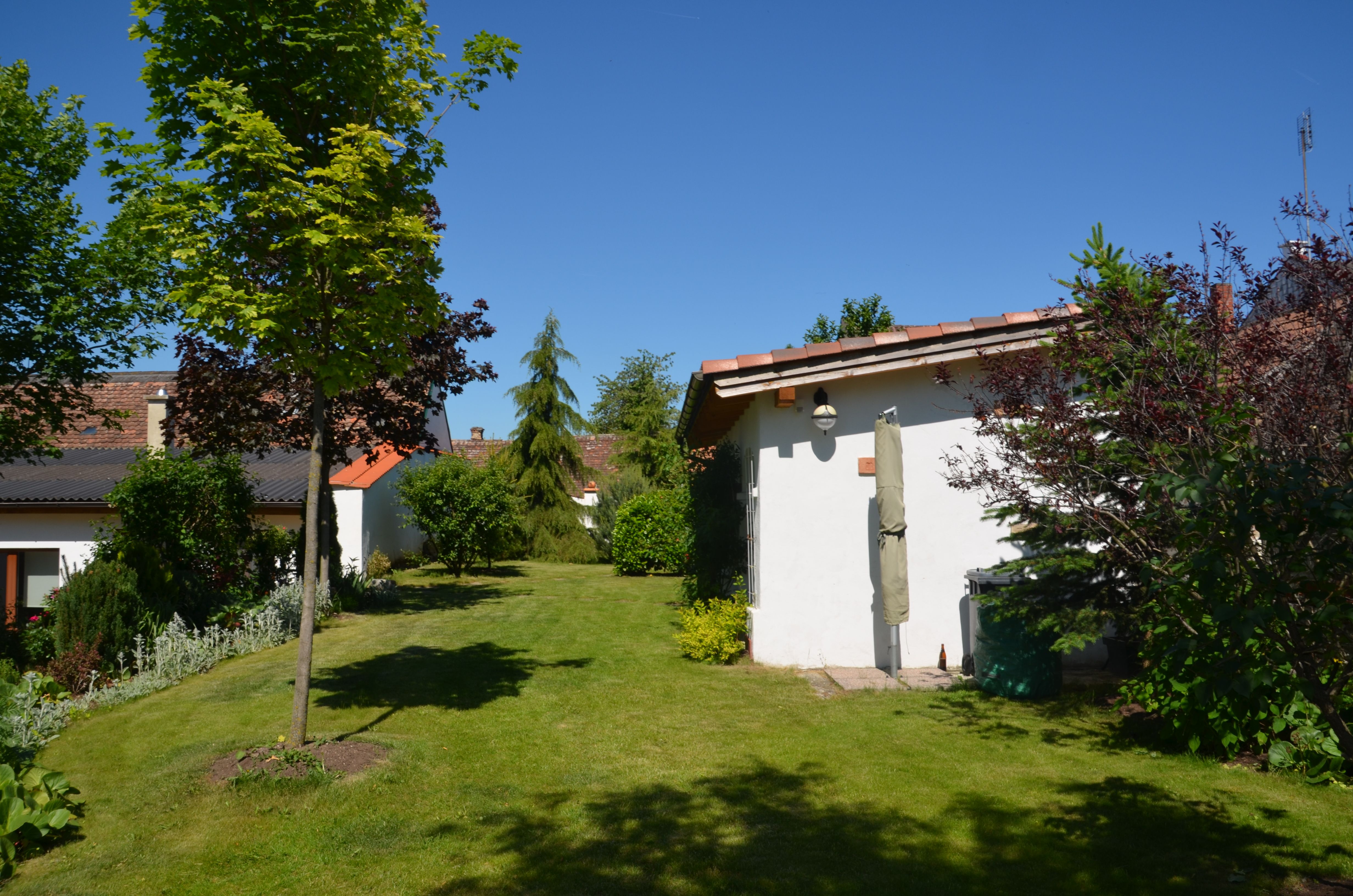 Ein gepflegter Garten mit Rasen, Bäumen und Sträuchern. Im Hintergrund sind zwei kleine Gebäude mit roten Dächern zu sehen.